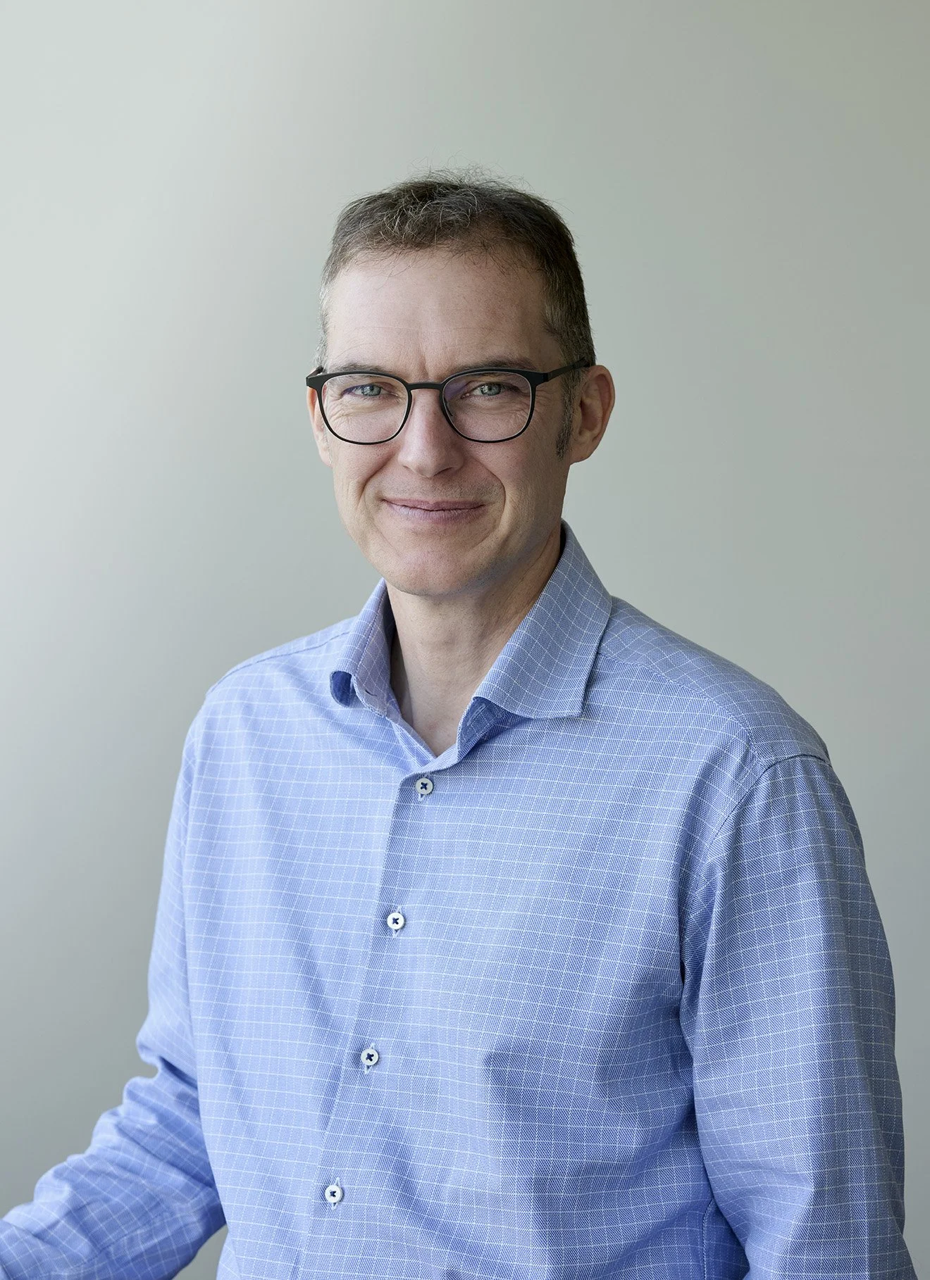 A man with short brown hair, glasses, and a light blue checkered shirt standing against a plain light-colored background.