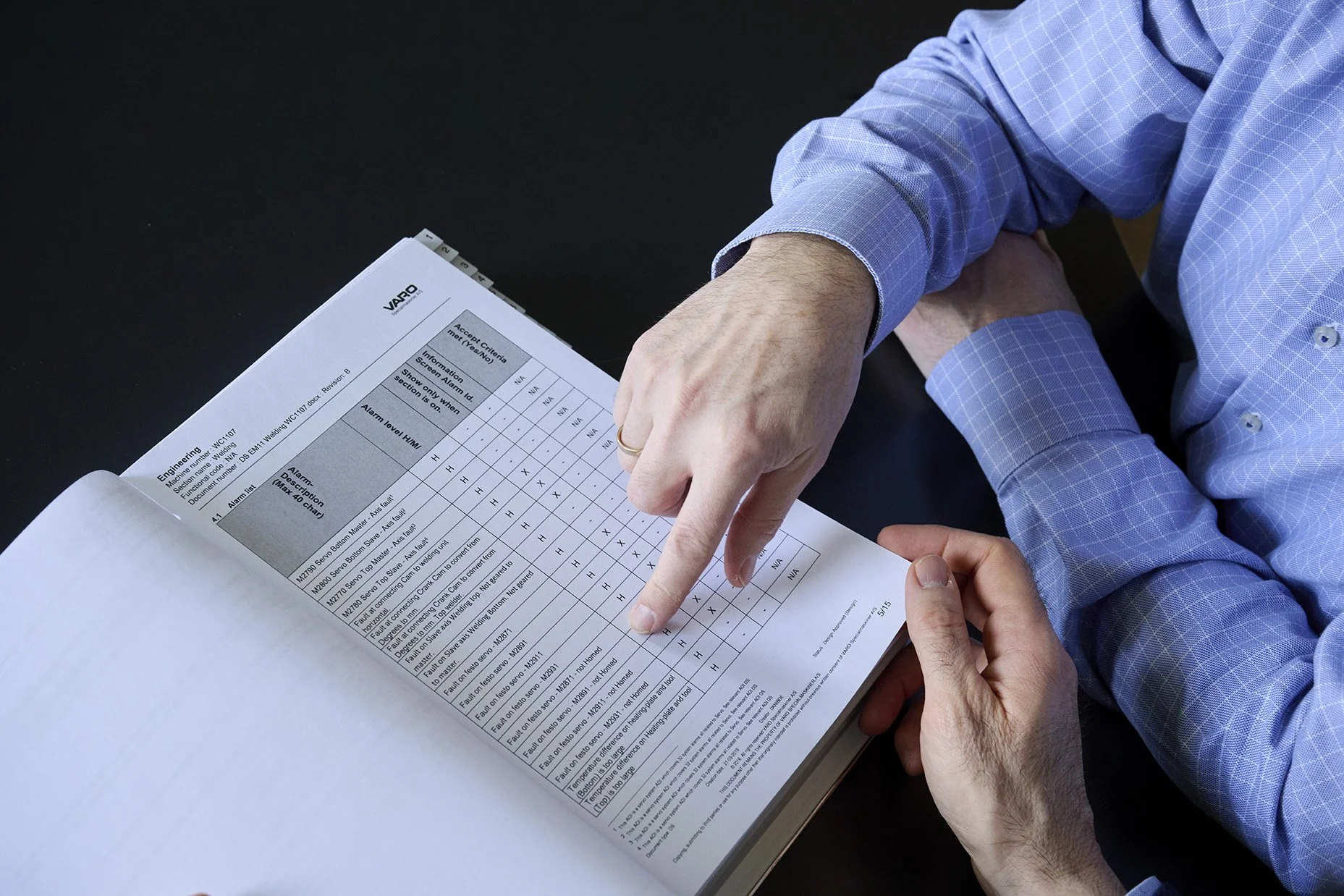 Two people, wearing blue checkered dress shirts, reviewing a printed technical document or chart with tabular data on a black surface.