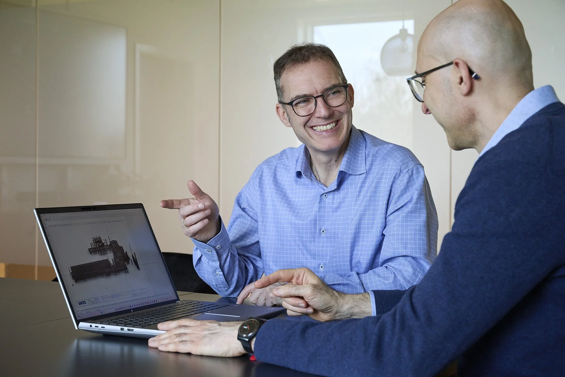 Two men engaged in a discussion at a table with a laptop displaying a technical drawing or 3D model. One man is smiling and gesturing with his hand, while the other man is listening attentively.
