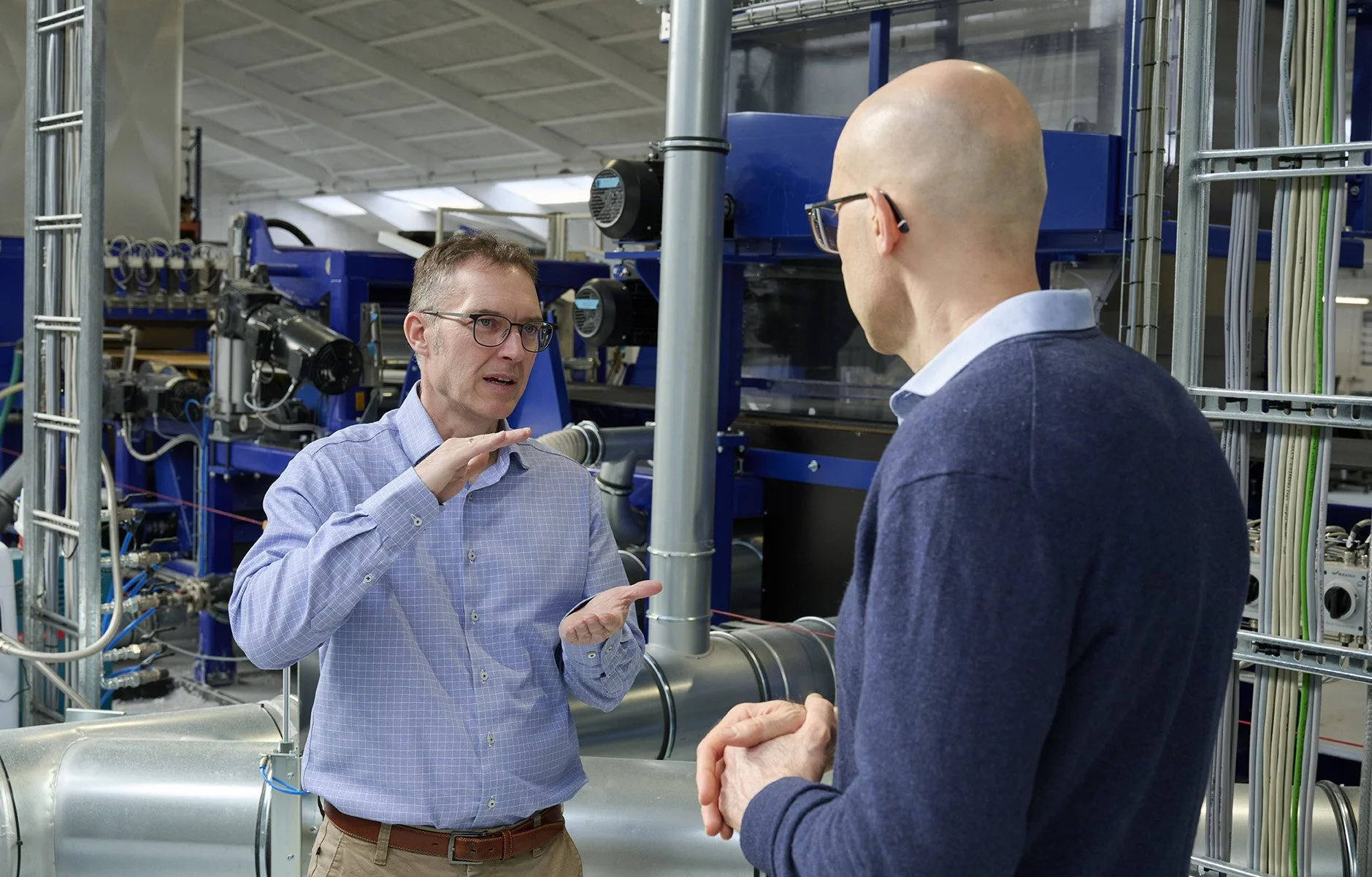 Two men in a technical discussion inside an industrial or laboratory environment with machinery and pipes around them.