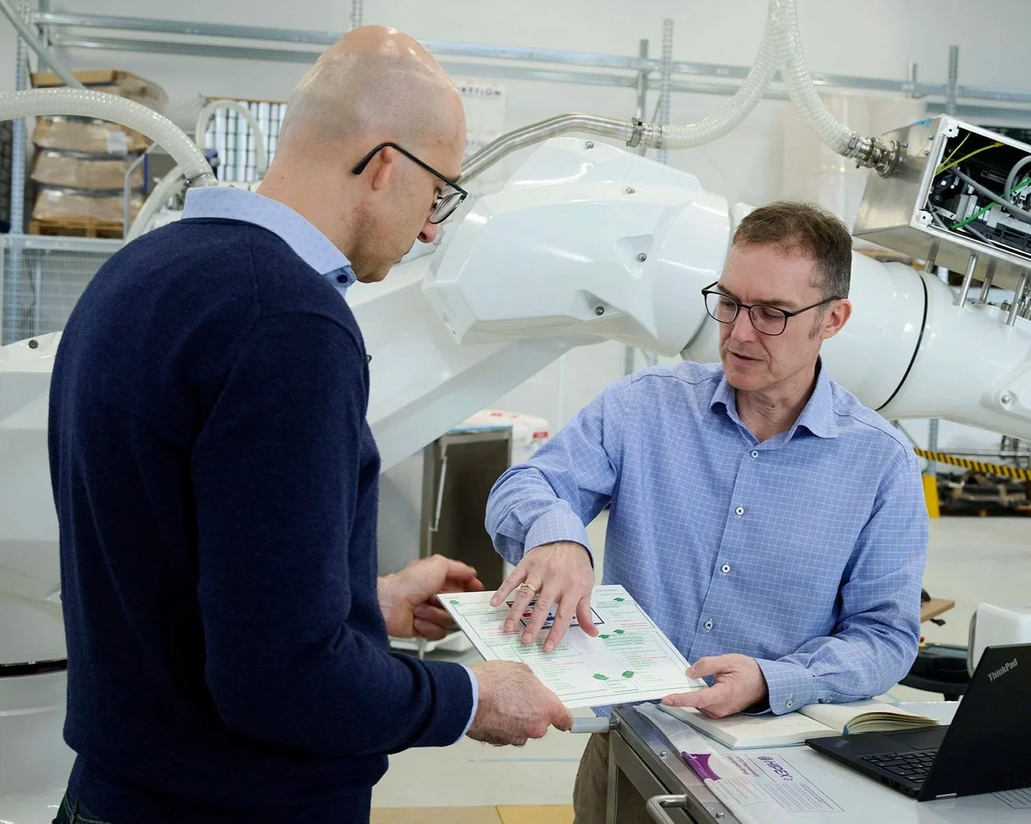 Two men in conversation in a laboratory or research facility, reviewing a document. One man is pointing at the document, while the other listens. There are large scientific machines and equipment in the background.