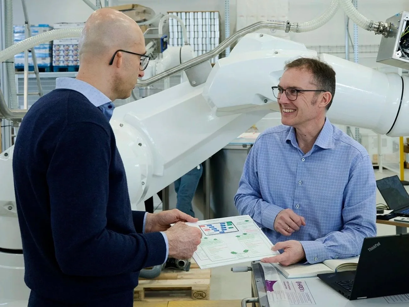 Two men in a lab or industrial setting discussing documents with charts. One man is holding papers, the other smiling. Large robotic arm in the background.