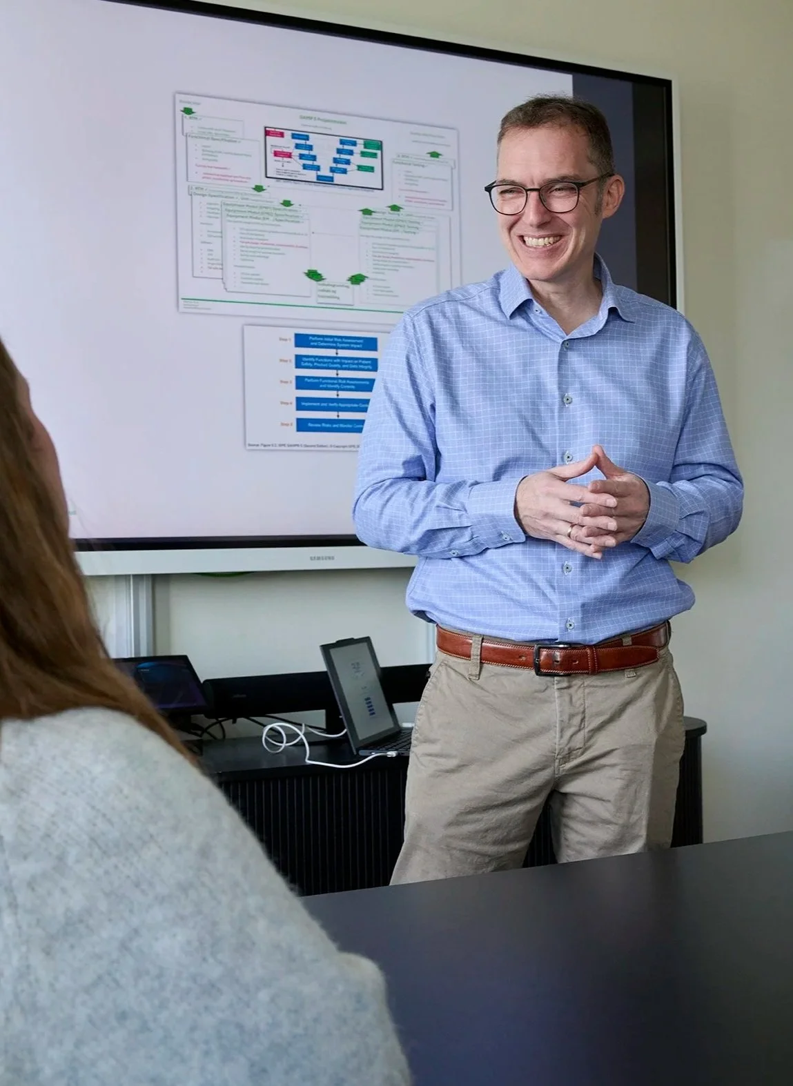 A man in glasses and a blue checkered shirt giving a presentation to a group, standing in front of a large screen displaying a flowchart or diagram.
