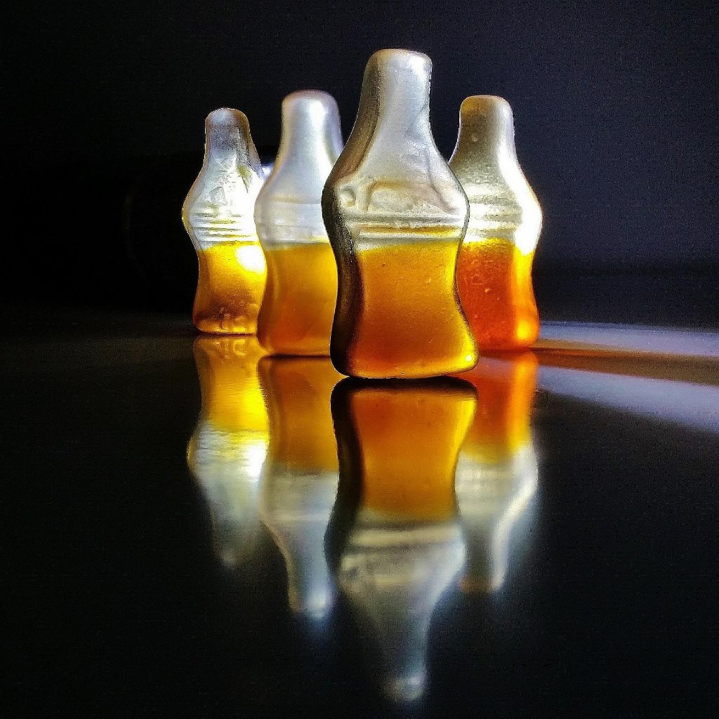 Four cola bottle candies in a line with a dark background, illuminated from behind, reflecting on a shiny surface.