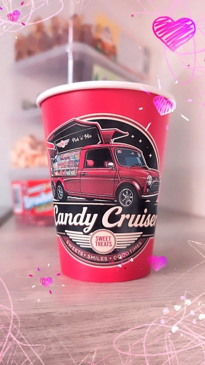 A pink paper cup with the Candy Cruiser logo on it. The cup also says "Pick n Mix" and "Sweet Treats" with decorative text and icons.