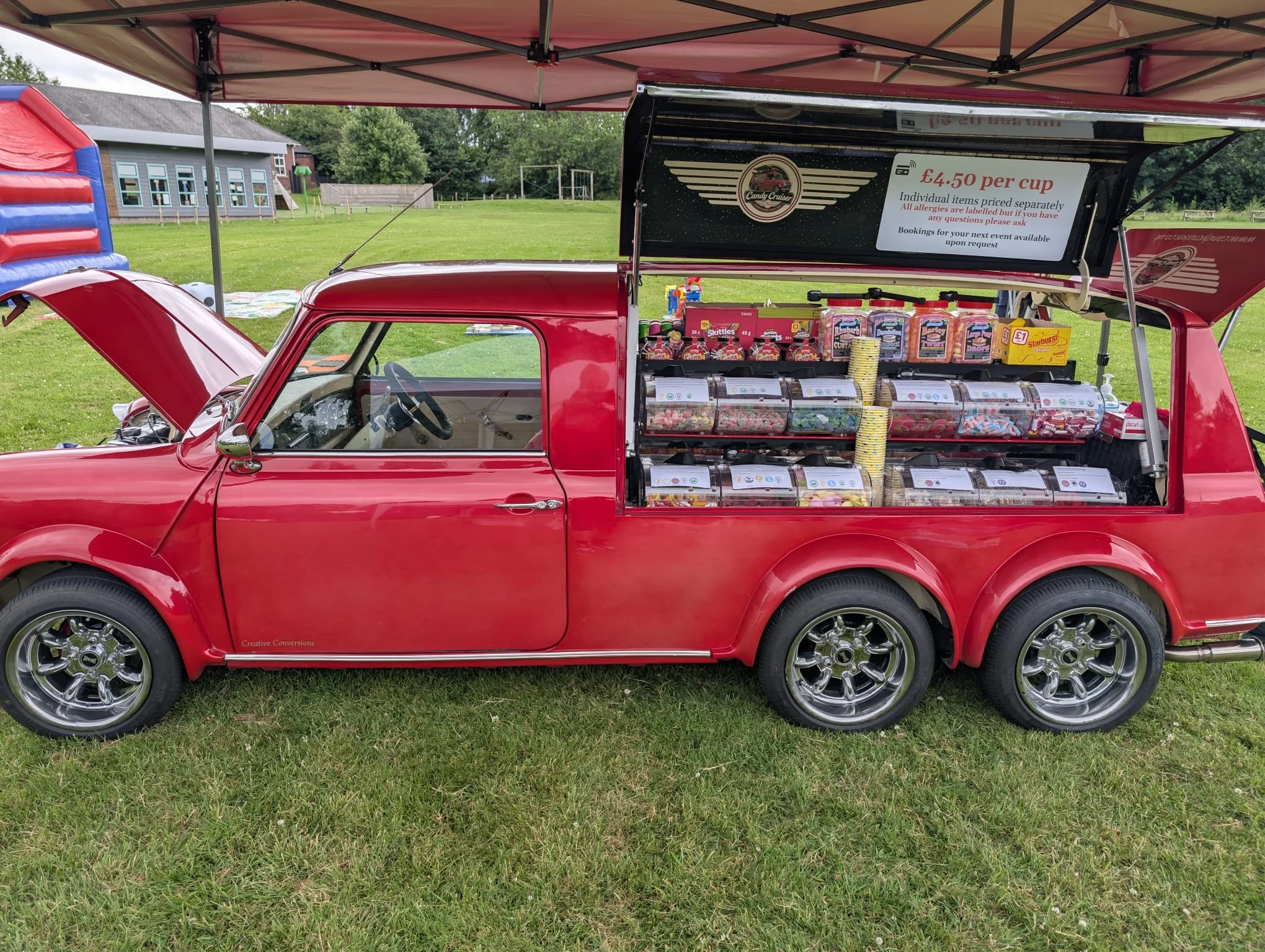 A red vintage car converted into a mobile candy stand, parked on a grassy field under a canopy. The car's side is open to display various candies and sweets, with a sign above advertising candy prices and booking details.