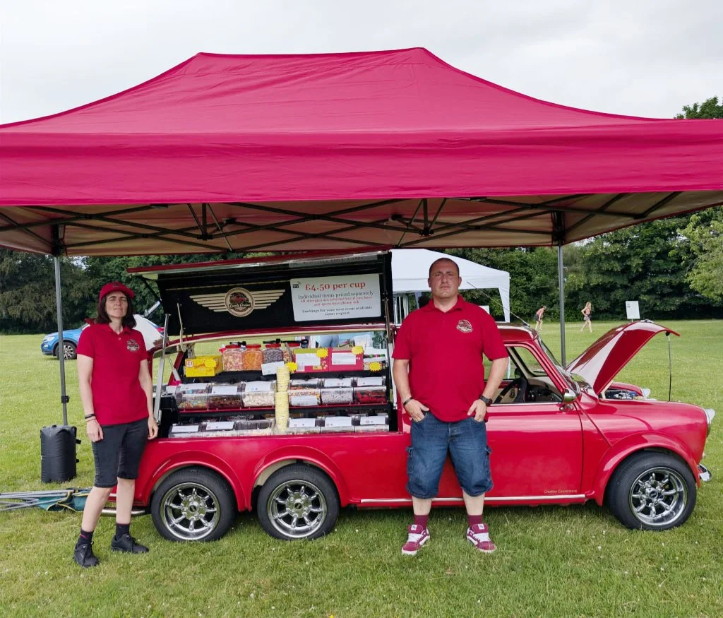 Two people standing in front of a vintage red car with an open gull-wing sides which display a variety of candies, under a red canopy tent in a park.