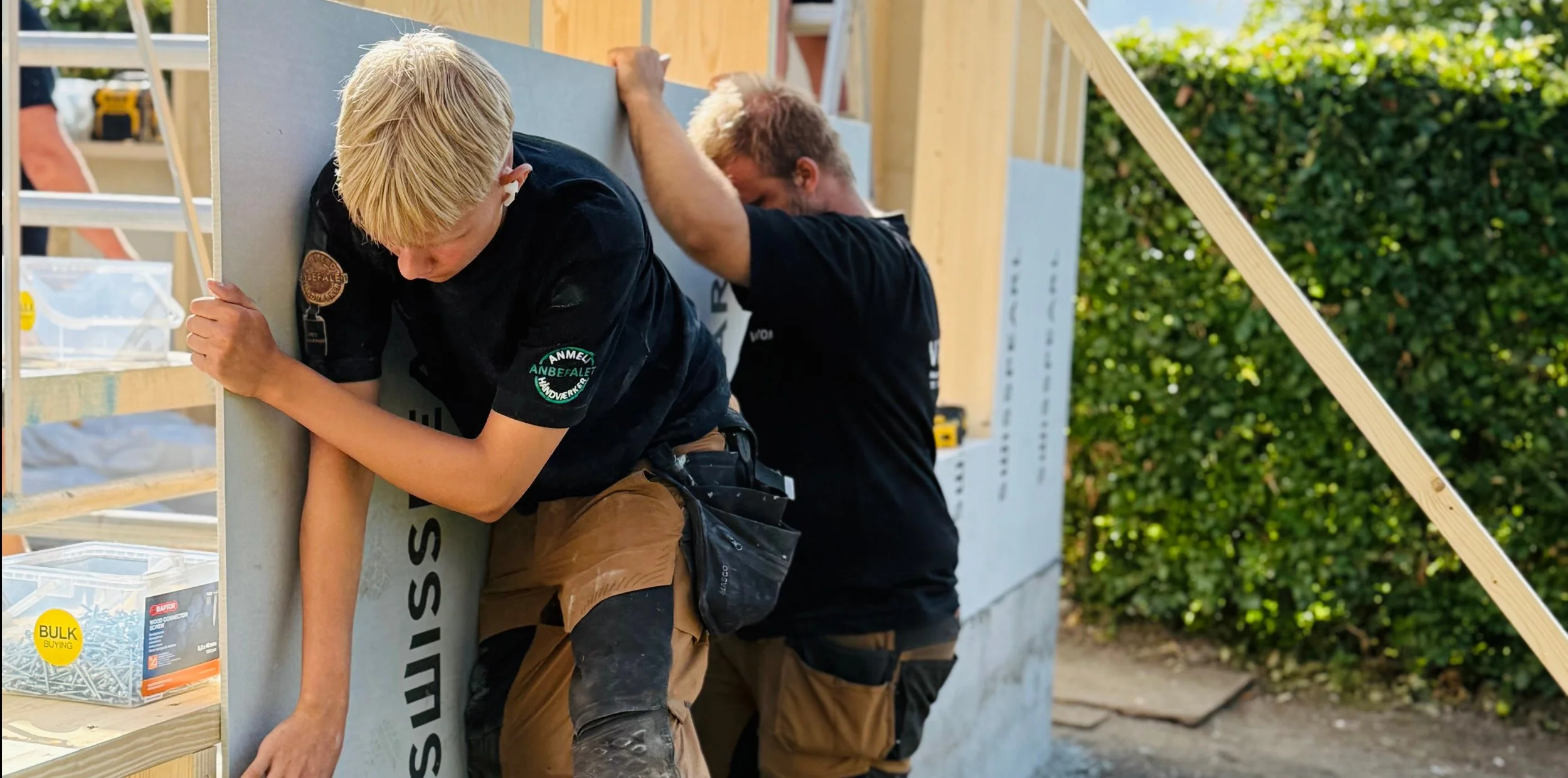 To mænd arbejder på en boligbyggeplads, hvor de installerer en væg. Begge er iført arbejdstøj og værktøjsseler, og den ene har blondt hår.