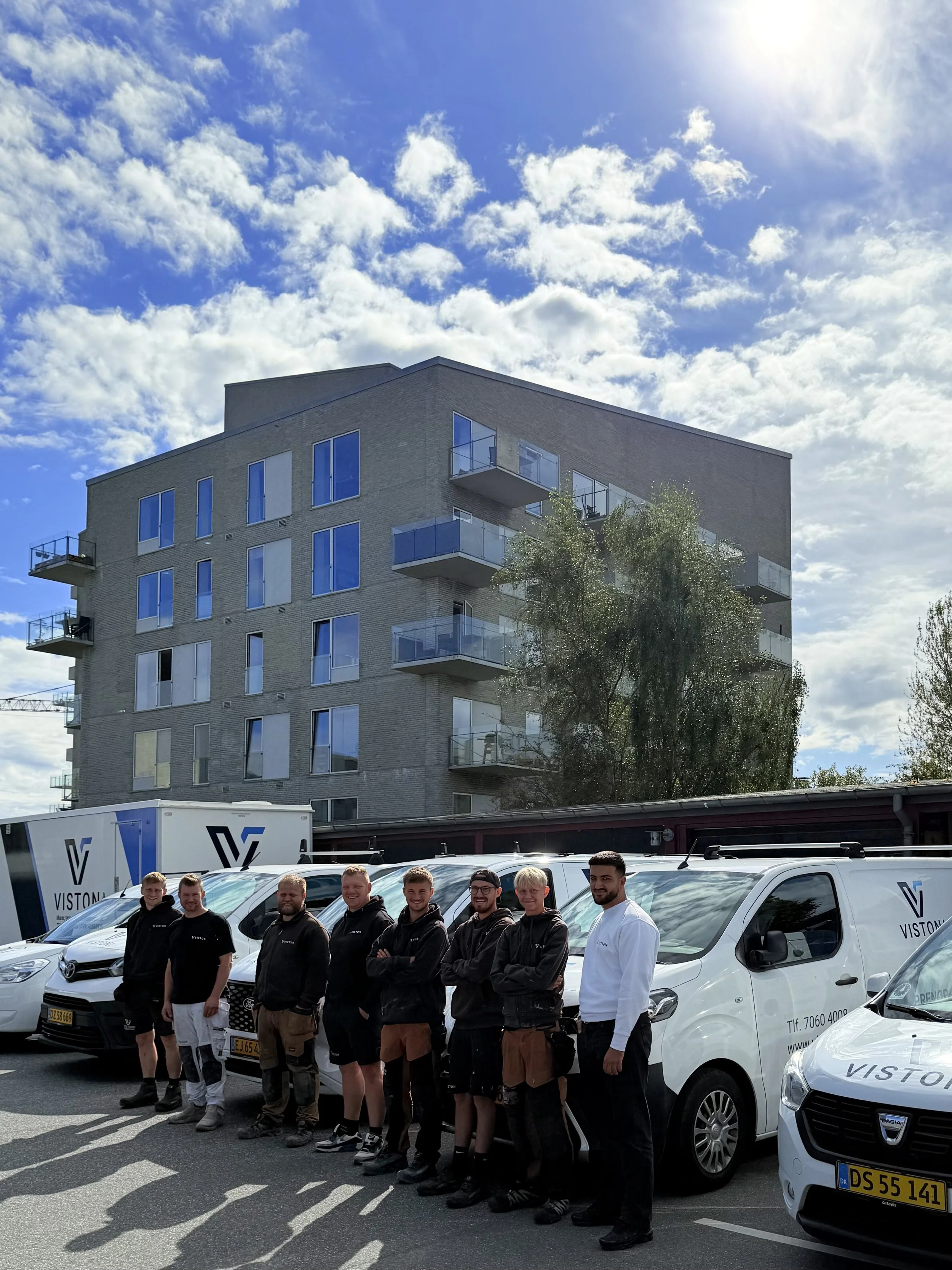 En gruppe mænd står foran hvide firmabiler, smilende, i en parkeringsplads med moderne boligblokke i baggrunden, under en skyet himmel.