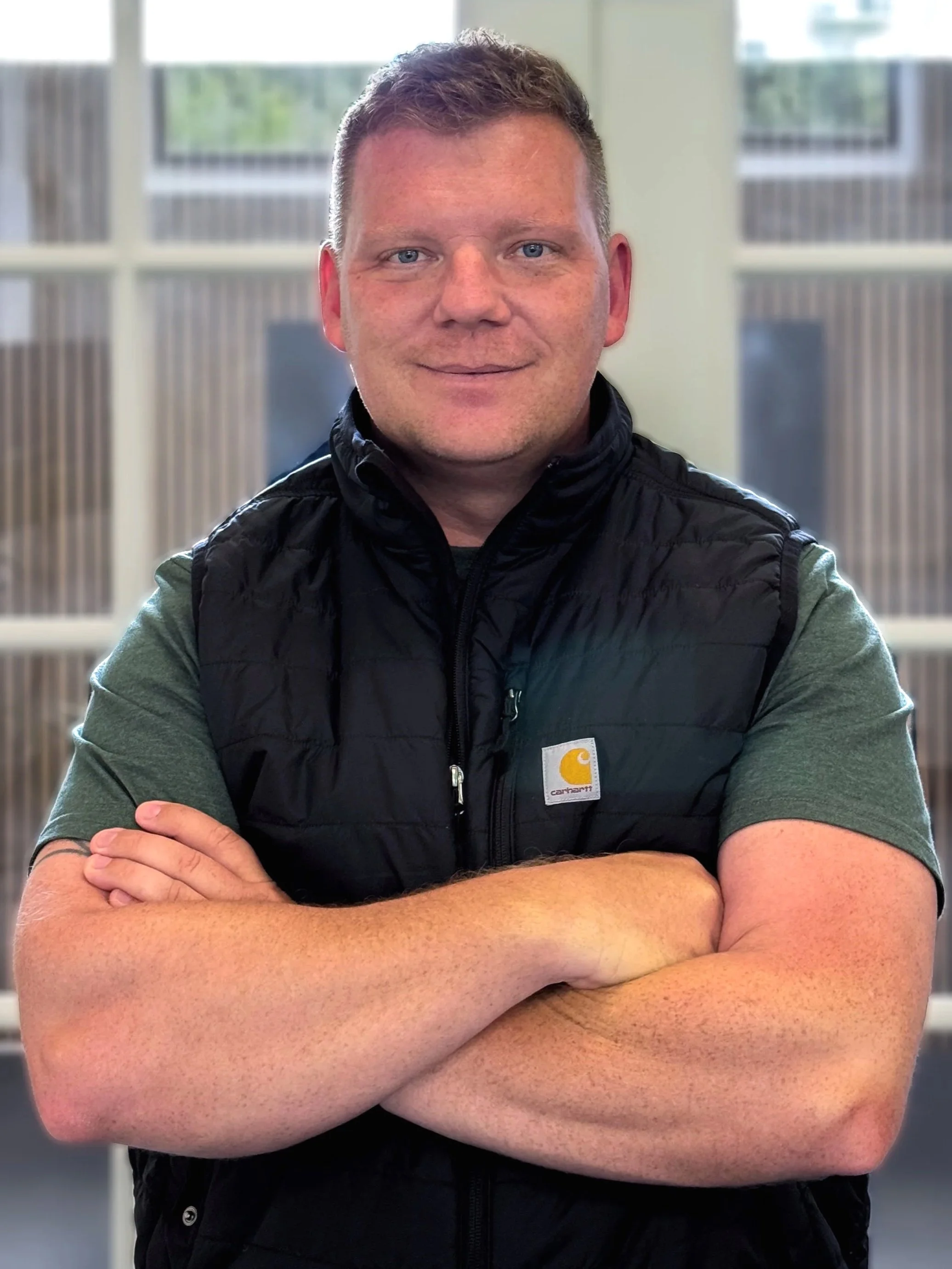 Et portræt af en mand med kort brunt hår, blå øjne, og lyst hud, iført en grøn T-shirt og en sort Carhartt vest, siddende med armene foldet foran sig i et rum med store vinduer i baggrunden.