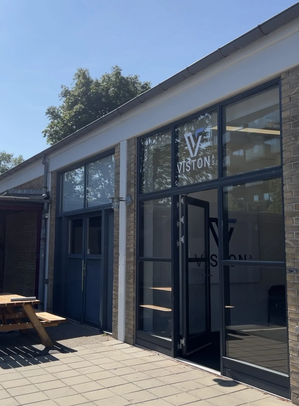 Et fritids- eller kontorlokale med store vinduer, der har logoet VISTON A/S på glasset. Der er en udendørs siddeplads med træbænke og et plettegulv foran bygningen.
