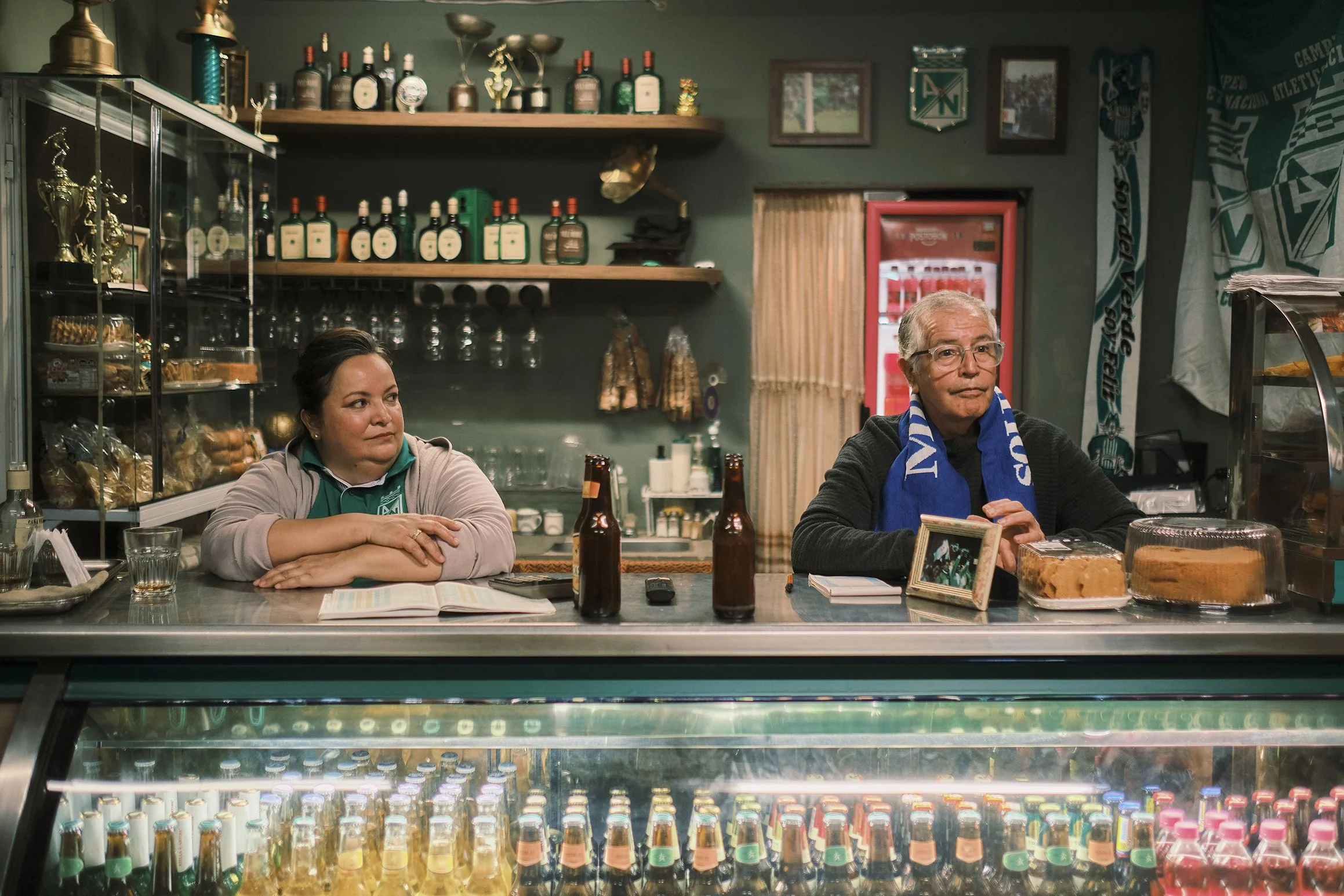 Dos personas, una mujer y un hombre, están detrás de un mostrador en un bar o cafetería, con bebidas y pasteles en exhibición, y decoraciones y botellas en el fondo.