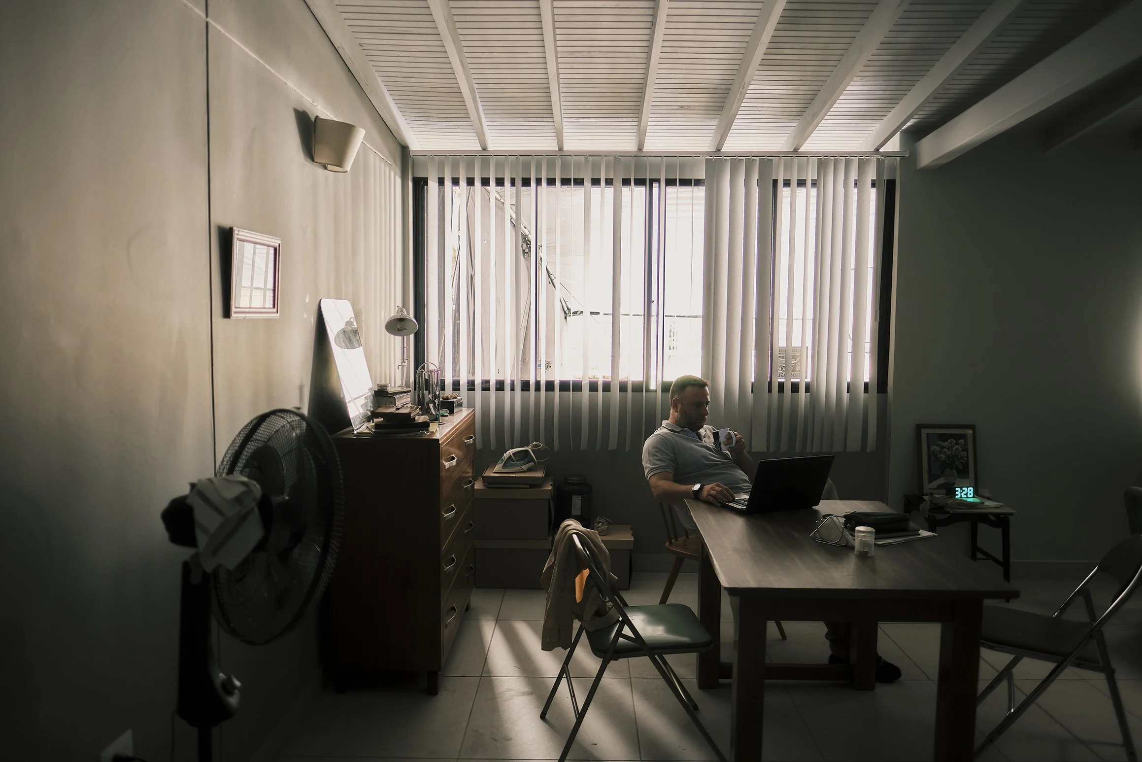 Oficina con un hombre sentado en la mesa, usando laptop y teléfono, luz natural entrando por ventanas con cortinas verticales.