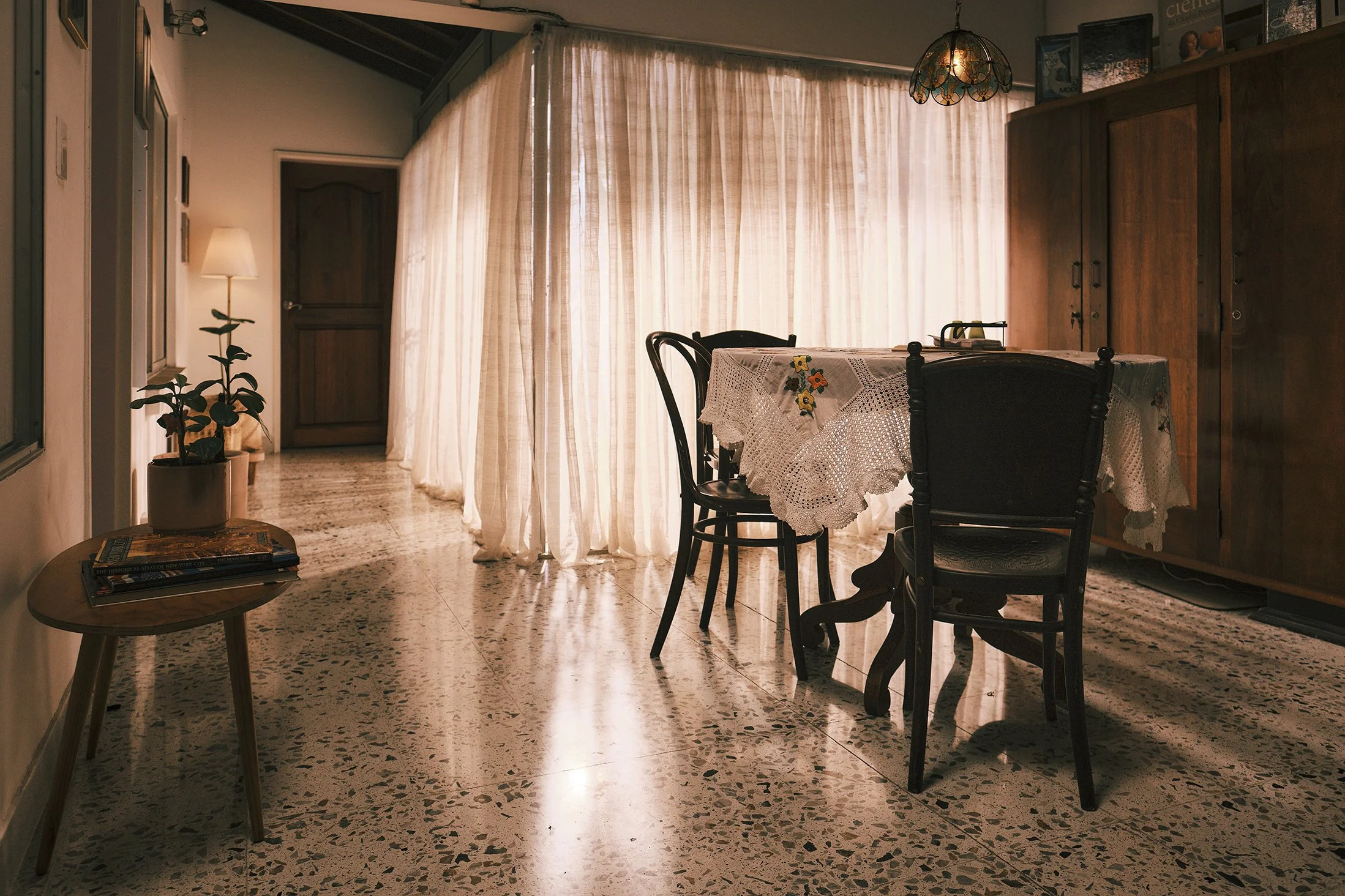 Interior de una sala con cortinas beige, una mesa con mantelería de encaje y sillas de madera, un armario de madera y una lámpara colgante de diseño artístico.