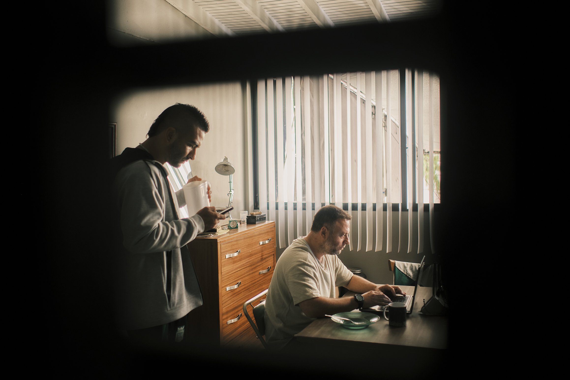 Dos hombres en una oficina, uno sentado en una mesa trabajando en una laptop y otro de pie revisando su teléfono, en un cuarto con cortinas verticales y luz natural