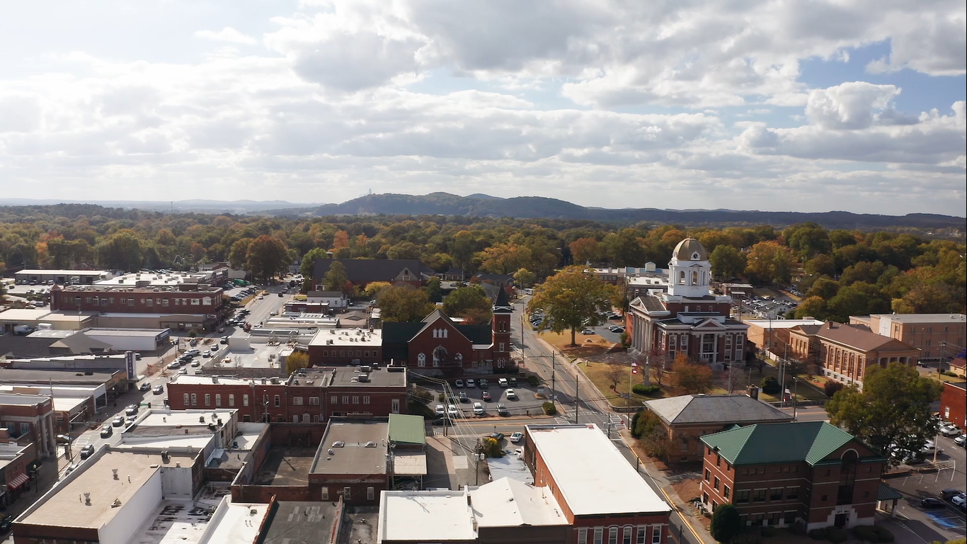 Aerial view of downtown small town with historic buildings, church, courthouse, parking lots, and surrounding trees with fall foliage, rolling hills in the distance, under partly cloudy sky.