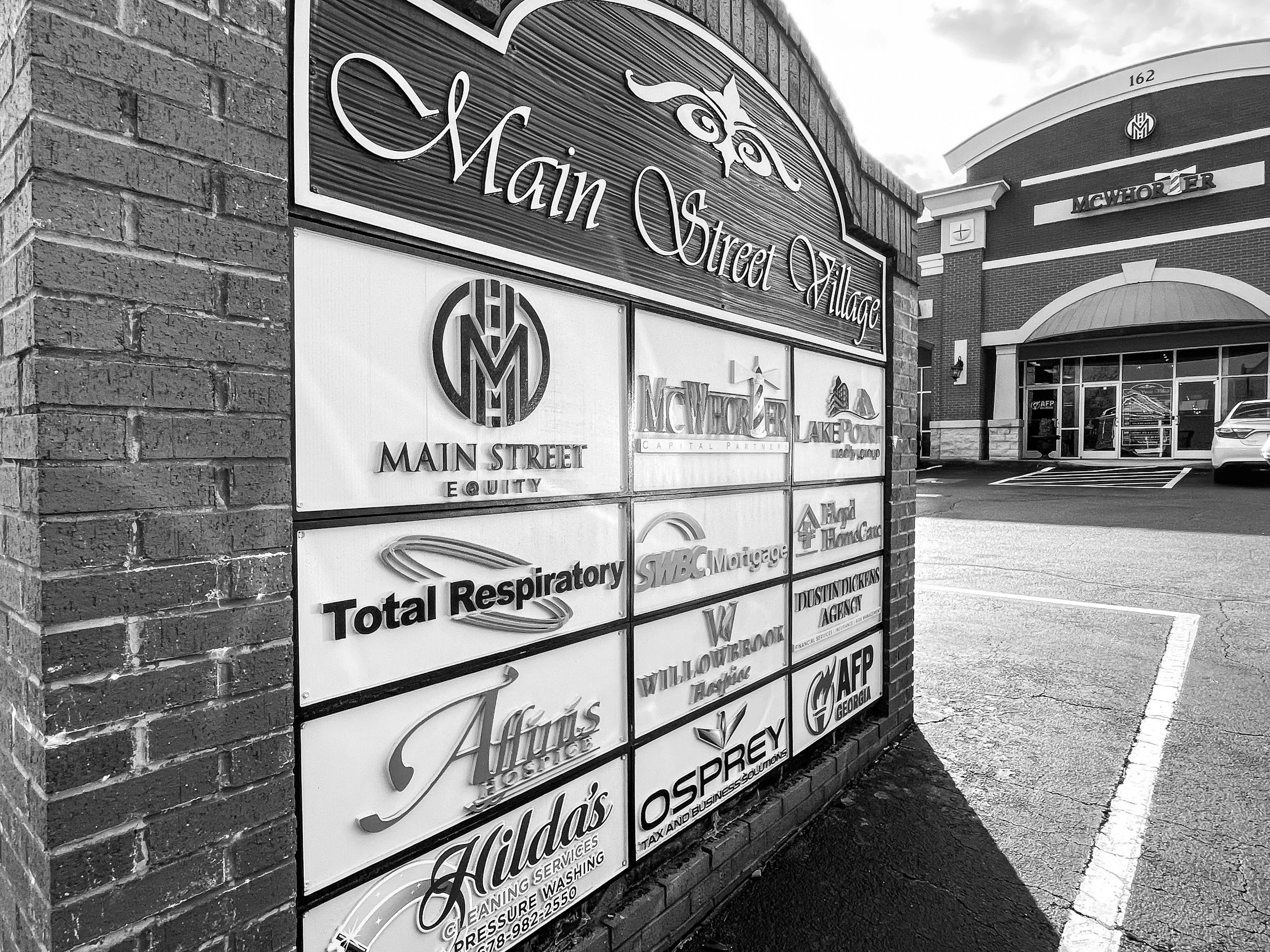 A large sign on a brick wall promoting businesses in Main Street Village. The sign lists various services including a furniture store, HVAC, flooring, plumbing, and more, with a shopping center entrance visible in the background.
