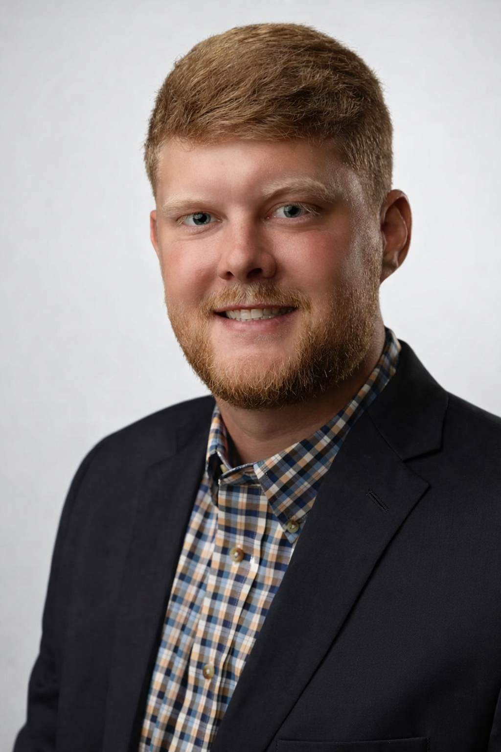 Headshot of a young man with red hair and a beard, wearing a dark blazer over a checkered shirt, smiling against a plain white background.