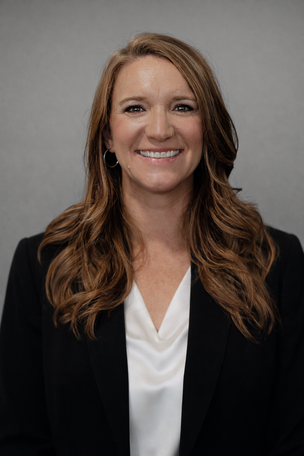 A woman with long wavy auburn hair, wearing a black blazer and a white camisole, smiling against a plain gray background.