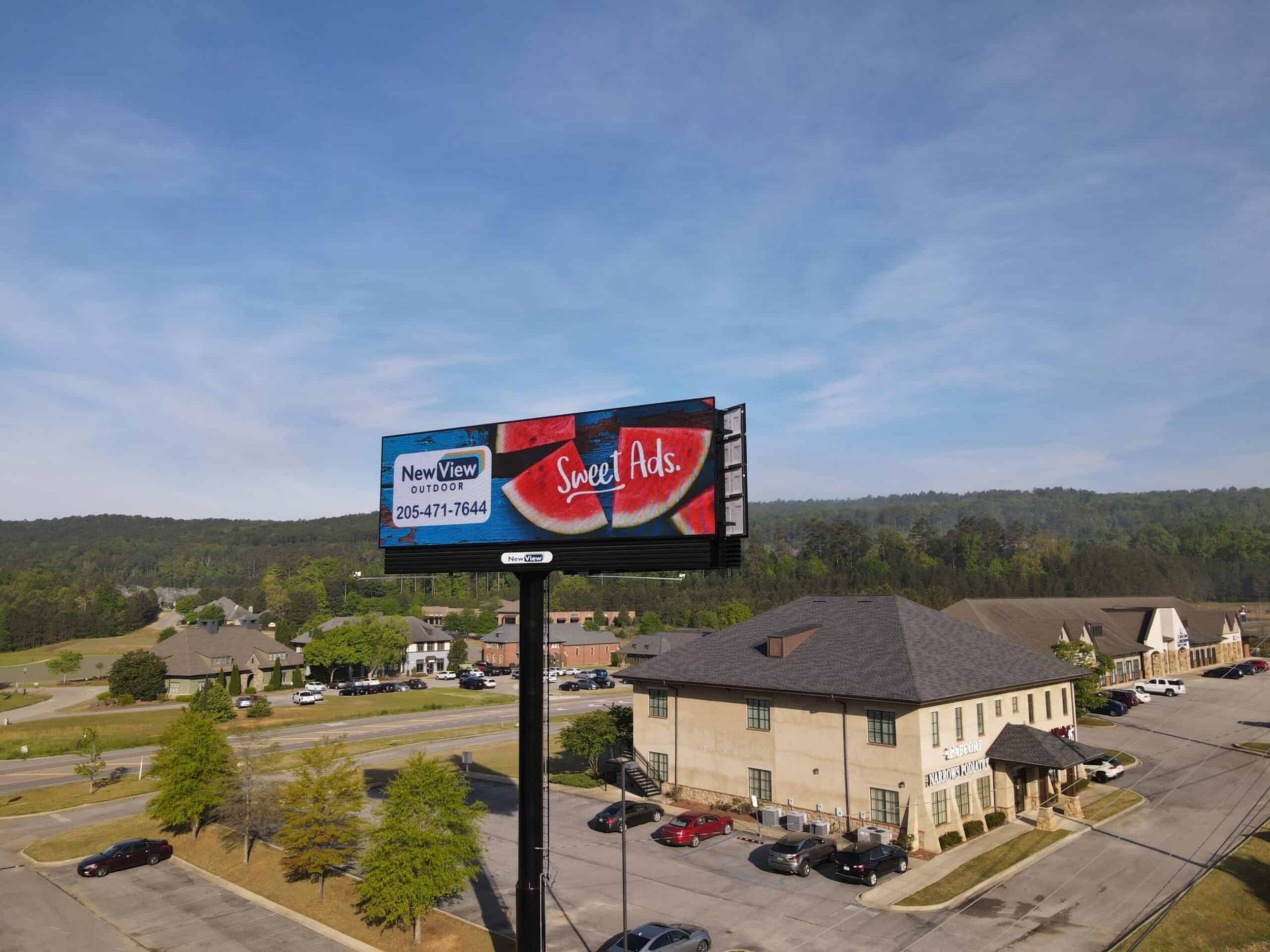 A large billboard advertising Sweet Ads watermelon slices, located over a parking lot and residential houses with green trees and a hilly landscape in the background, under a partly cloudy sky.