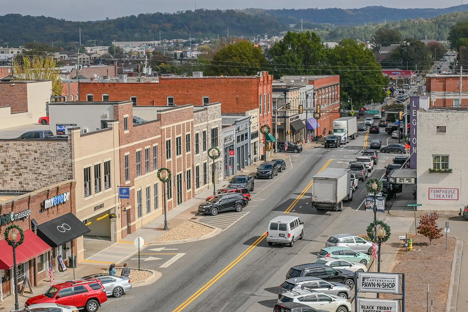 Downtown street view with brick and stone buildings, parked cars, moving vehicles, festive wreaths on lampposts, a theater sign, and a hilly background with trees and distant hills.