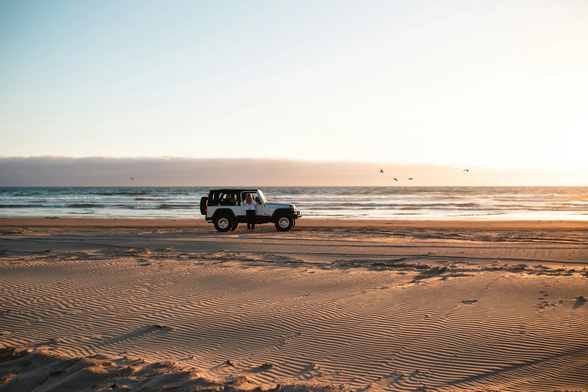 A person standing outside a white Jeep vehicle parked on a beach during sunset, with the ocean and sky in the background, and birds flying in the distance.