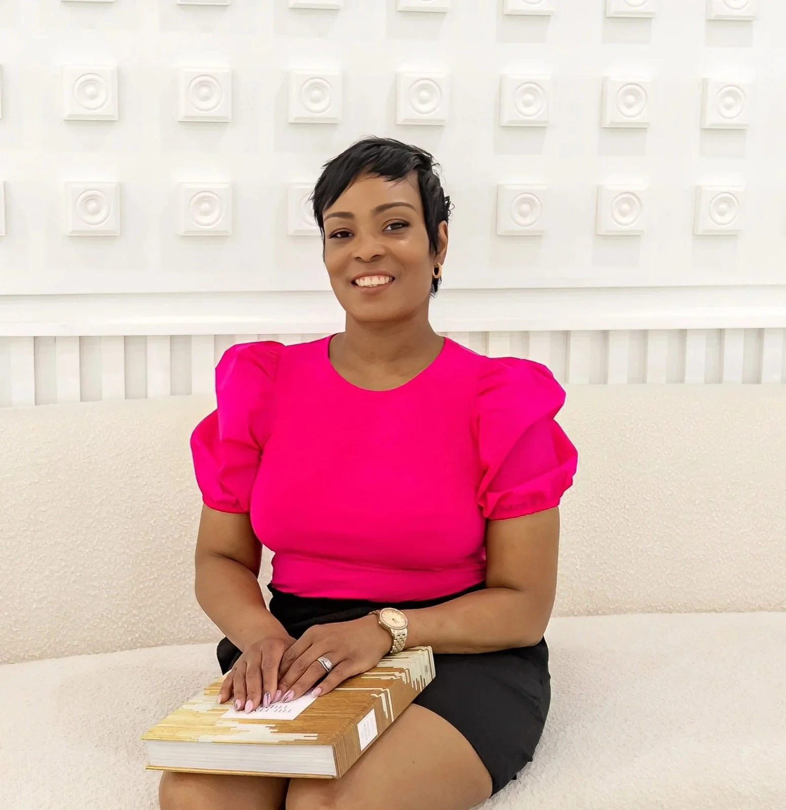 A woman with short black hair, wearing a bright pink blouse and black skirt, sitting on a light-colored sofa, holding a closed book, and smiling at the camera.