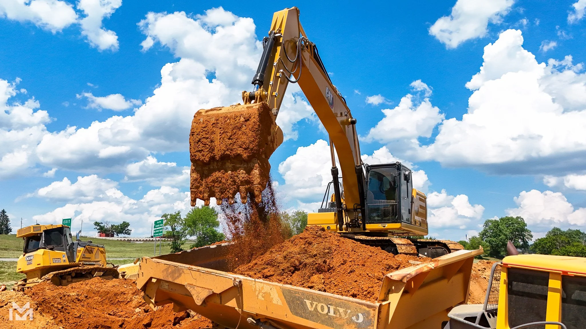 A large yellow excavator lifting a load of dirt against a bright blue sky with white clouds, working on a construction site.