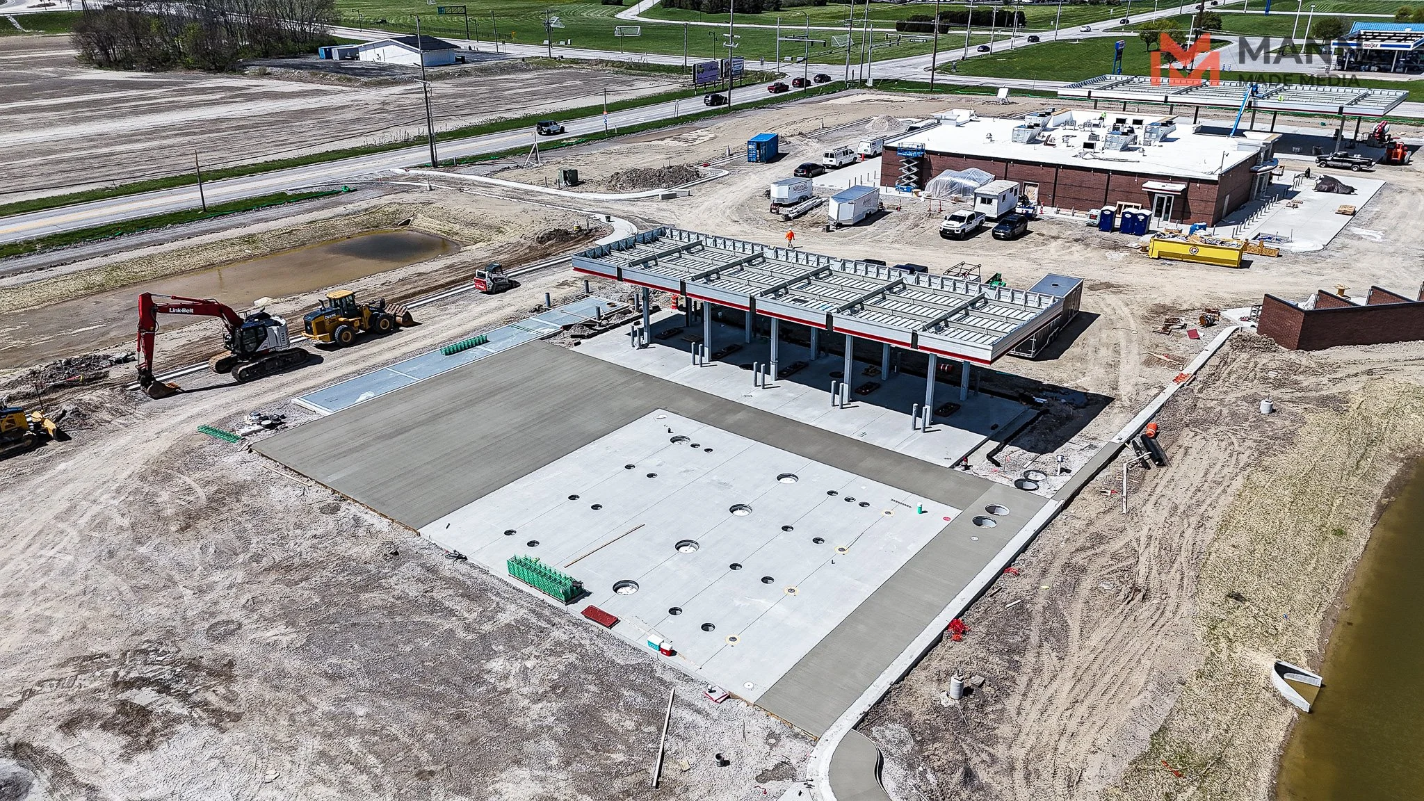 An aerial view of a construction site where a new building is being constructed. The foundation and some concrete slabs are completed, and construction machinery and workers are visible on site. Nearby, there is a gas station with multiple fuel pumps.