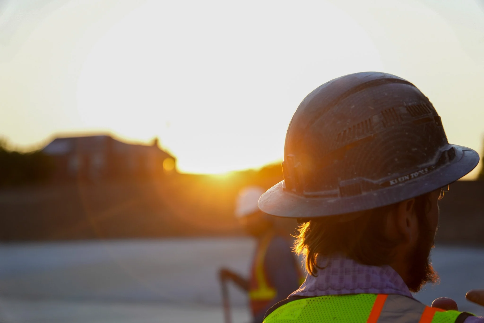 Person wearing a safety helmet and reflective vest, standing outdoors at sunset.