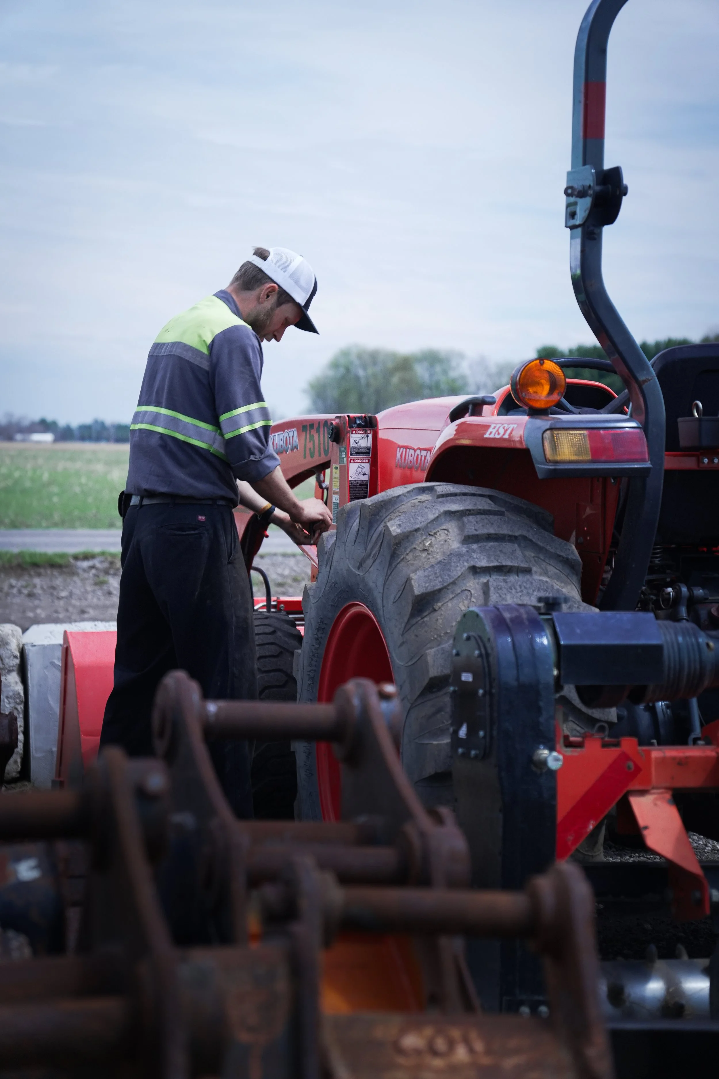 A man wearing a cap and work uniform is repairing or inspecting a red Kubota tractor outdoors, with fields and trees in the background.