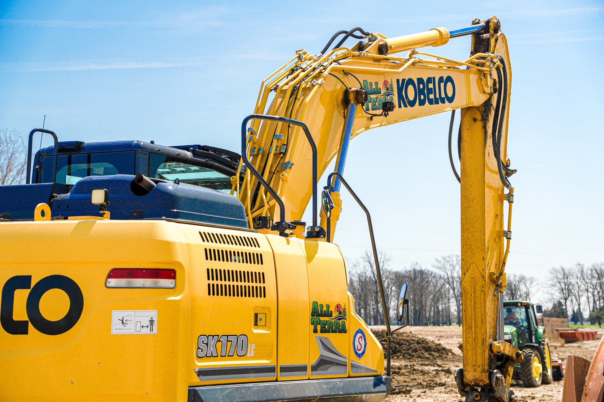 Close-up of a yellow excavator with the words 'All Terra' on the arm, operating at a construction site with a clear blue sky in the background.