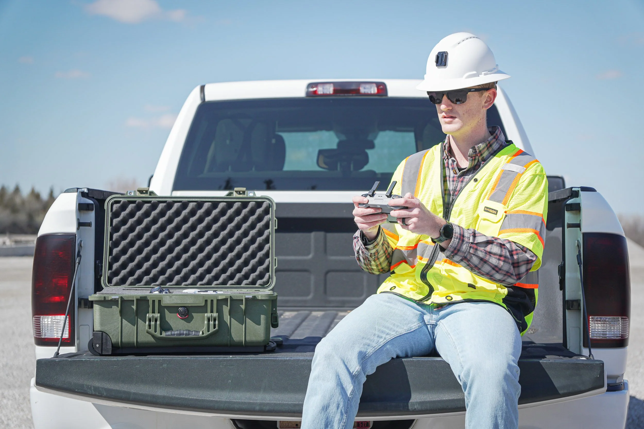 A construction worker in a yellow safety vest and white hard hat sitting on the tailgate of a white pickup truck, operating a drone controller with a large protective case nearby.