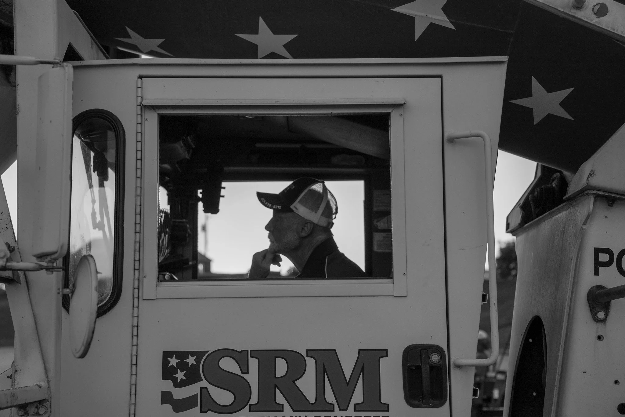 A man wearing a cap and sunglasses inside a concrete mixer truck, with part of a sign and a large star-spangled background visible in black and white.