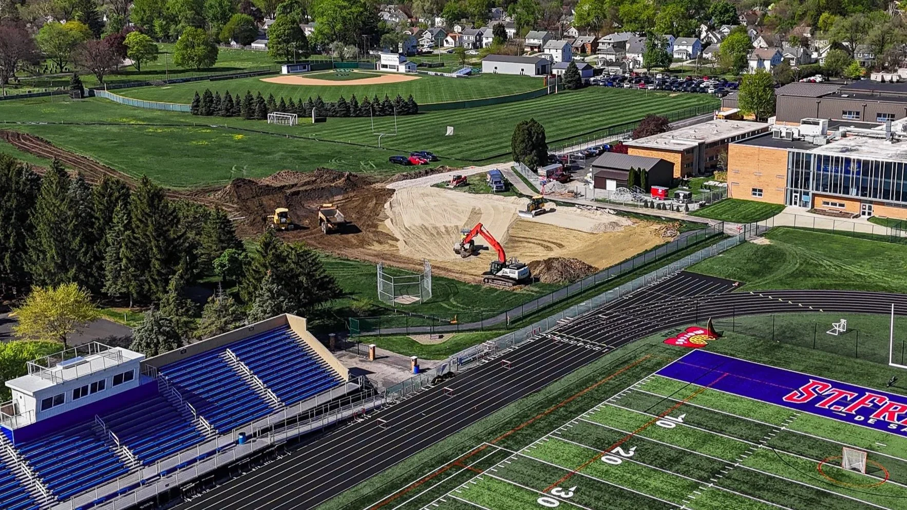 A baseball stadium with rows of blue seats, a running track with a red and yellow star logo, and a large athletic field. Behind the stadium, there is ongoing construction with excavators, dirt piles, and construction vehicles, including a backhoe. Further back, there is a sports field with a baseball diamond and a football field with visible goalposts surrounded by a park and residential neighborhood.