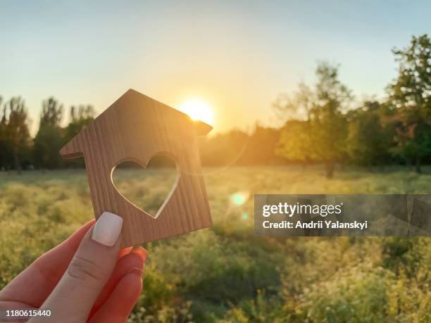 Hand holding a small wooden house with a heart-shaped cutout, sunset in the background over a field with trees.