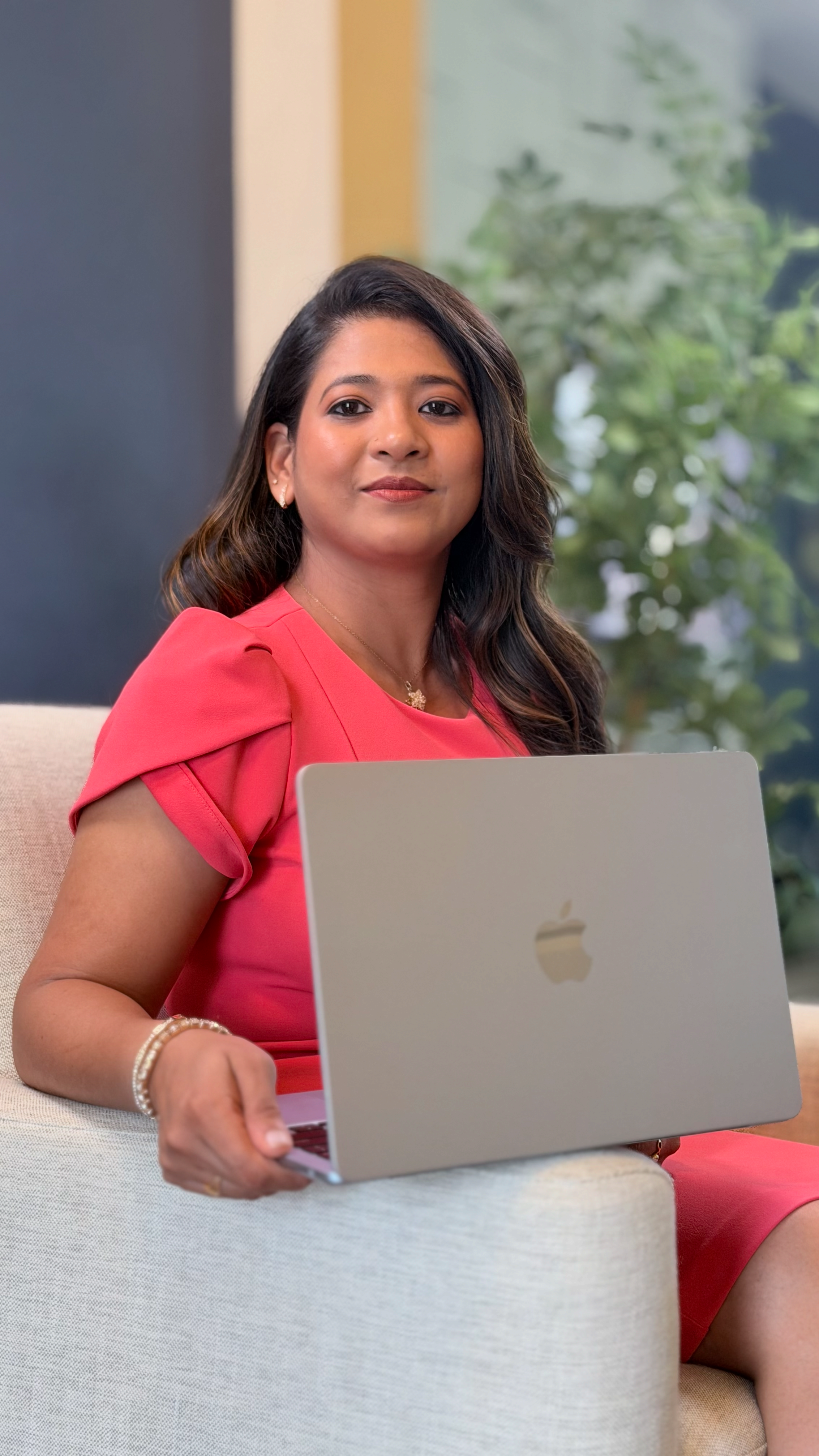 A woman with dark hair, wearing a pink dress and jewelry, sitting on a beige sofa in front of a green plant, holding a silver Apple MacBook laptop.