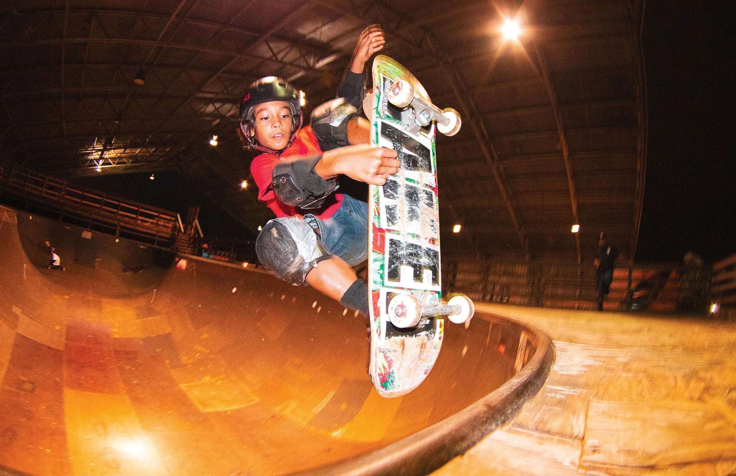 A young boy wearing a helmet and protective gear riding a skateboard inside a skatepark, with a curved ramp in the background and a large light overhead.