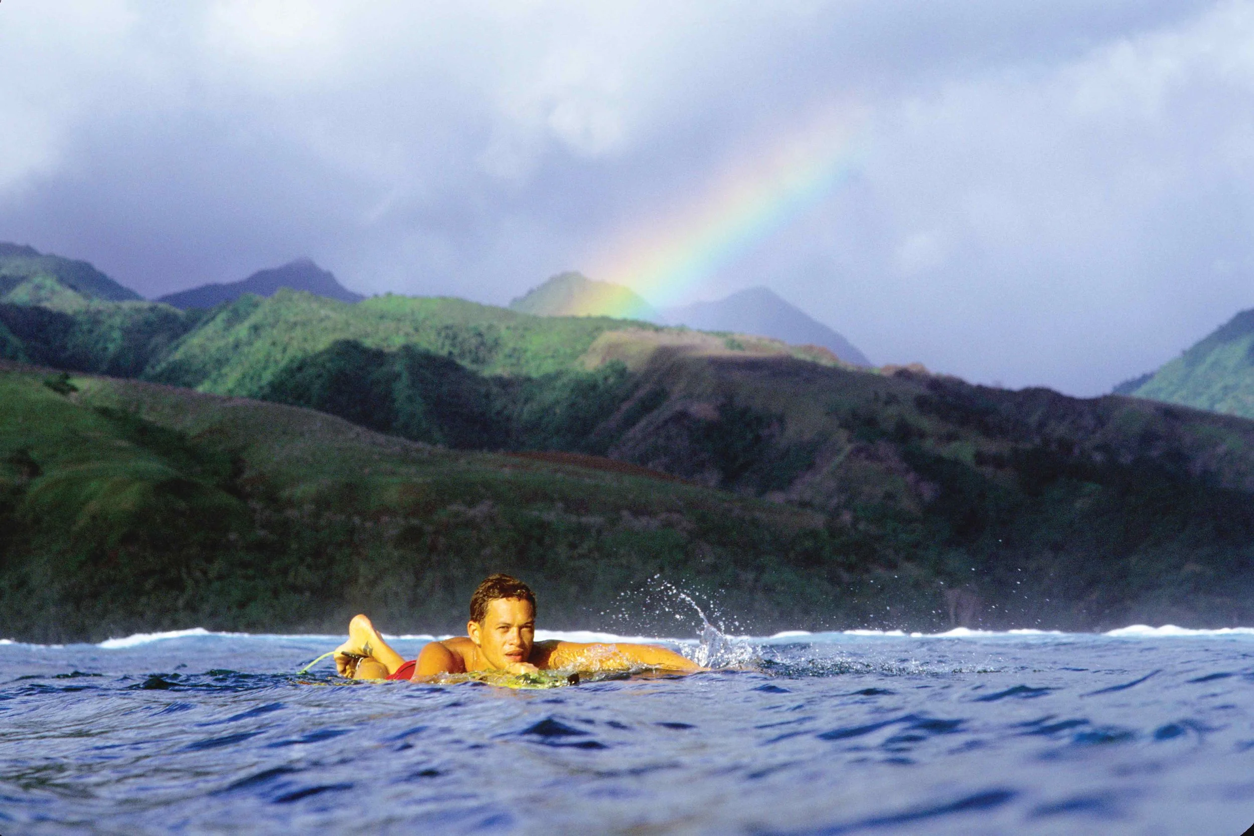 A man swimming in the ocean with lush green hills and a rainbow in the background.