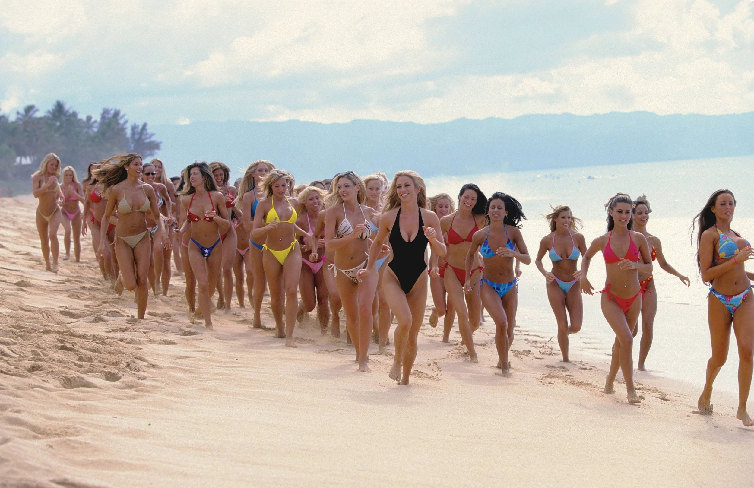 A group of women in swimsuits walking along a sandy beach with the ocean and sky in the background.