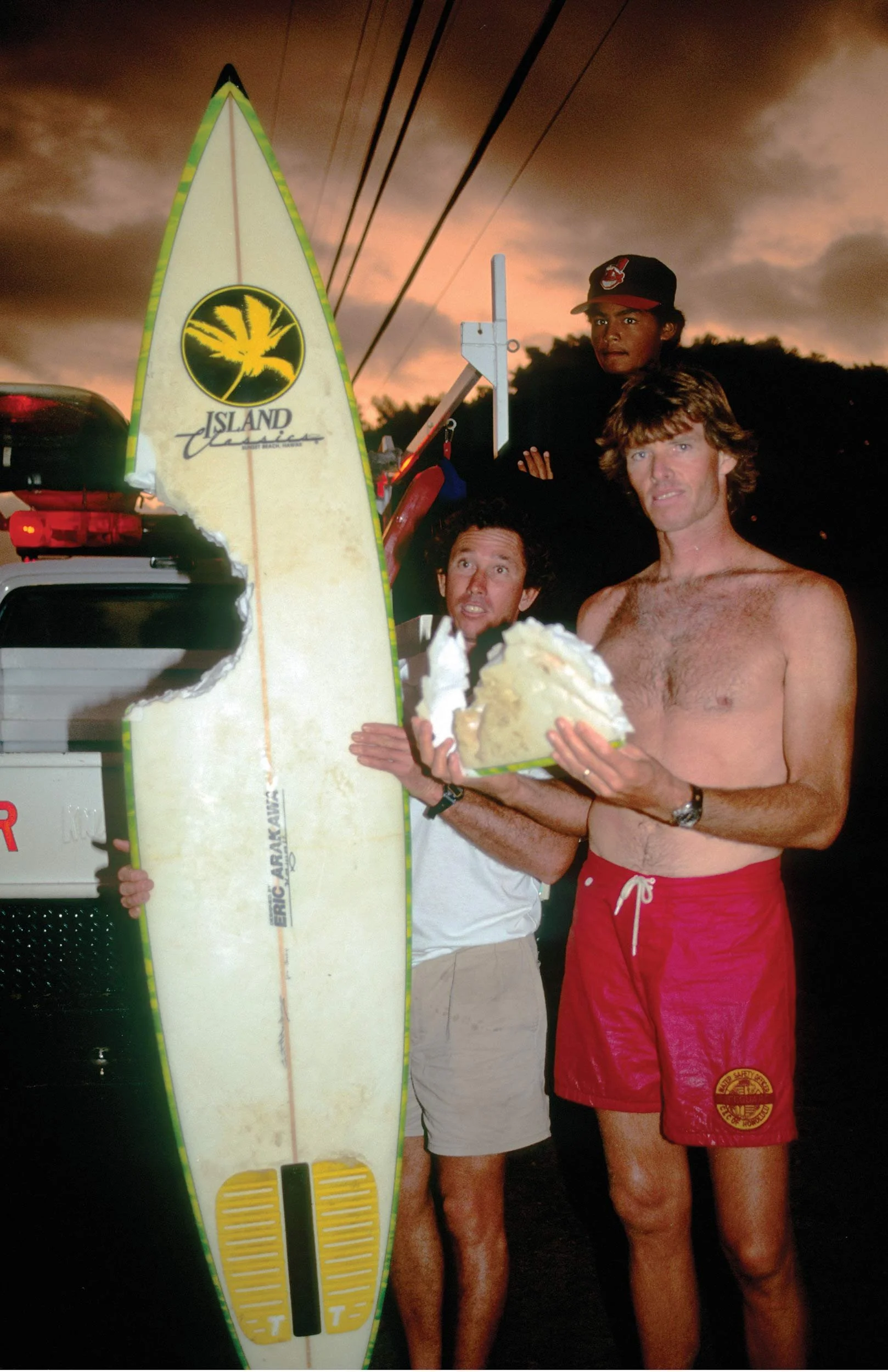 A group of shirtless men, some wearing red swim trunks and one with a black cap, stand outdoors at sunset. They are holding a large yellow and white surfboard with a logo that reads 'Island' and a black and yellow design. One man is holding a large p