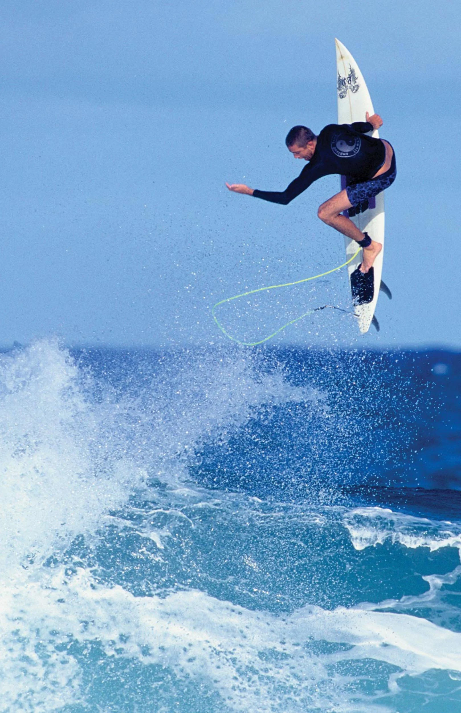 A man skydiving over the ocean, holding a surfboard, dressed in wetsuit and shorts, with water spray beneath him.