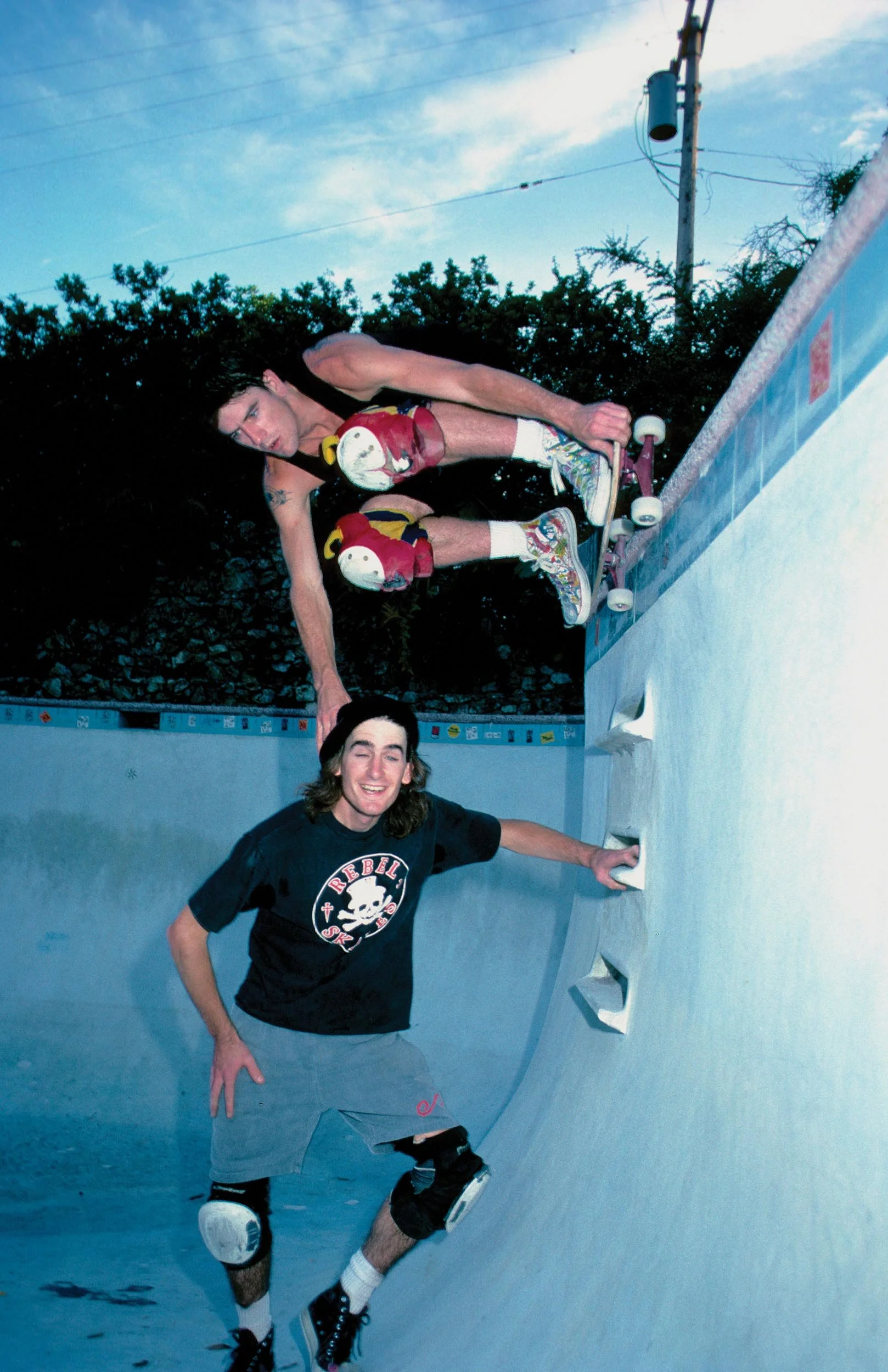 Three young men enjoying skating in a skate park with a concrete bowl, blue skies, and a utility pole in the background. One is skateboarding, another is smiling and posing, and the third is partially visible.