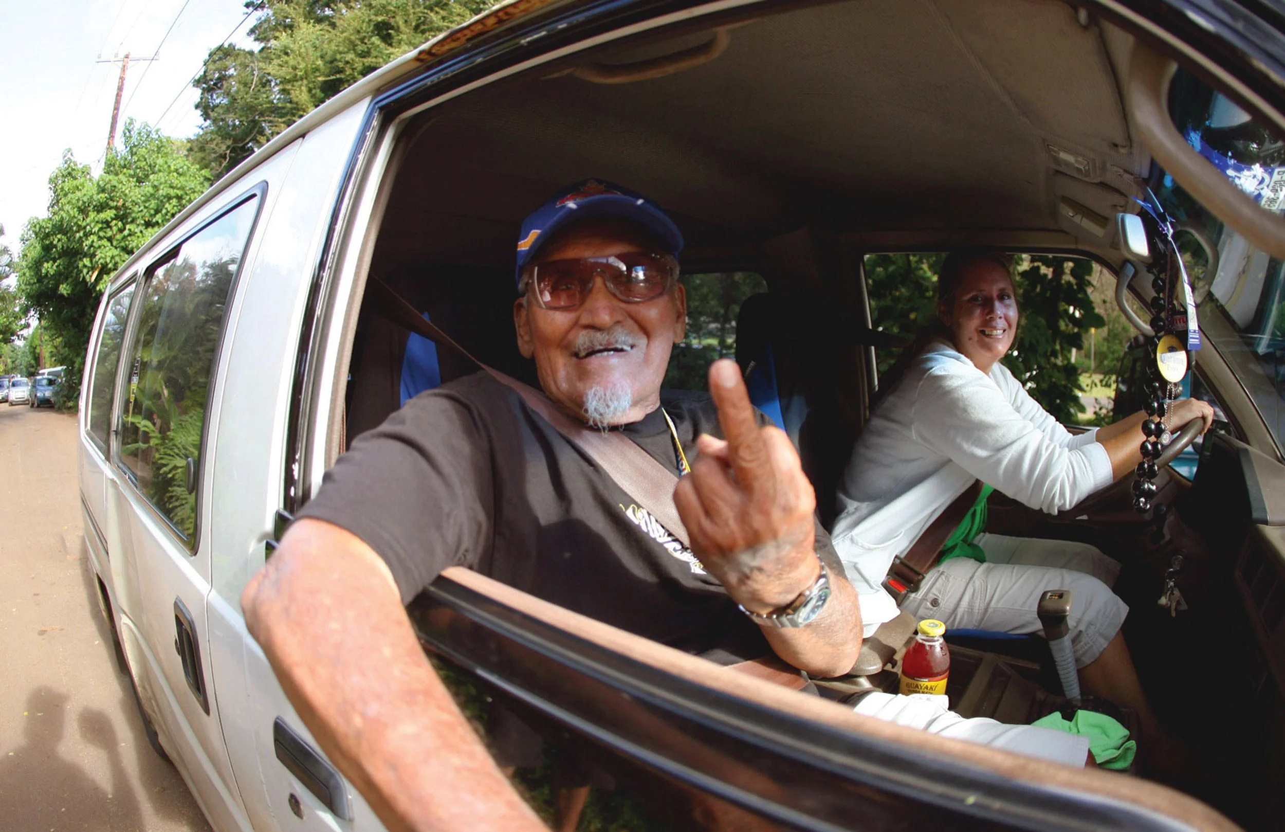 Two smiling people inside a white vehicle, one male wearing sunglasses and a cap giving a thumbs up, and one female driving, amidst green trees and parked cars.