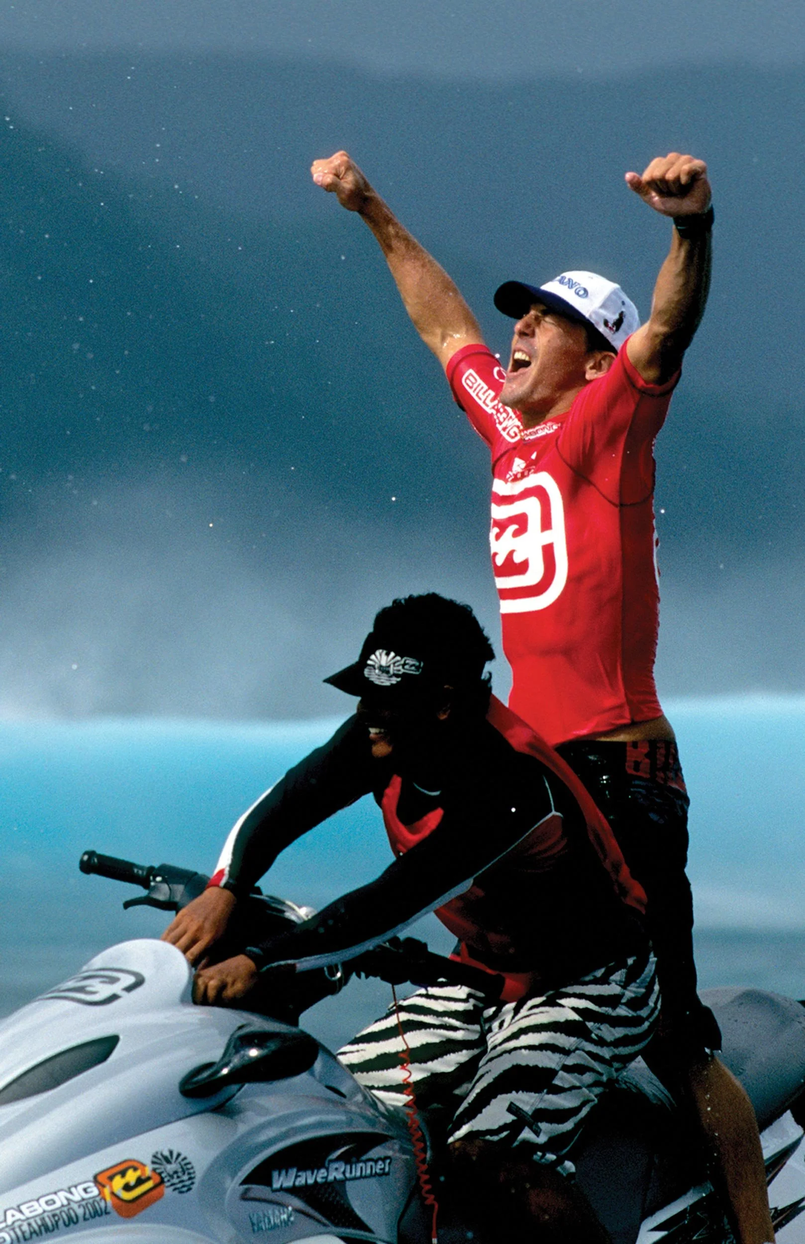 Two men riding a white jet ski on the water, one man standing on the jet ski with arms raised and wearing a red shirt and cap, the other man sitting and holding the handlebars, wearing striped shorts and a black jacket, during a cloudy day.
