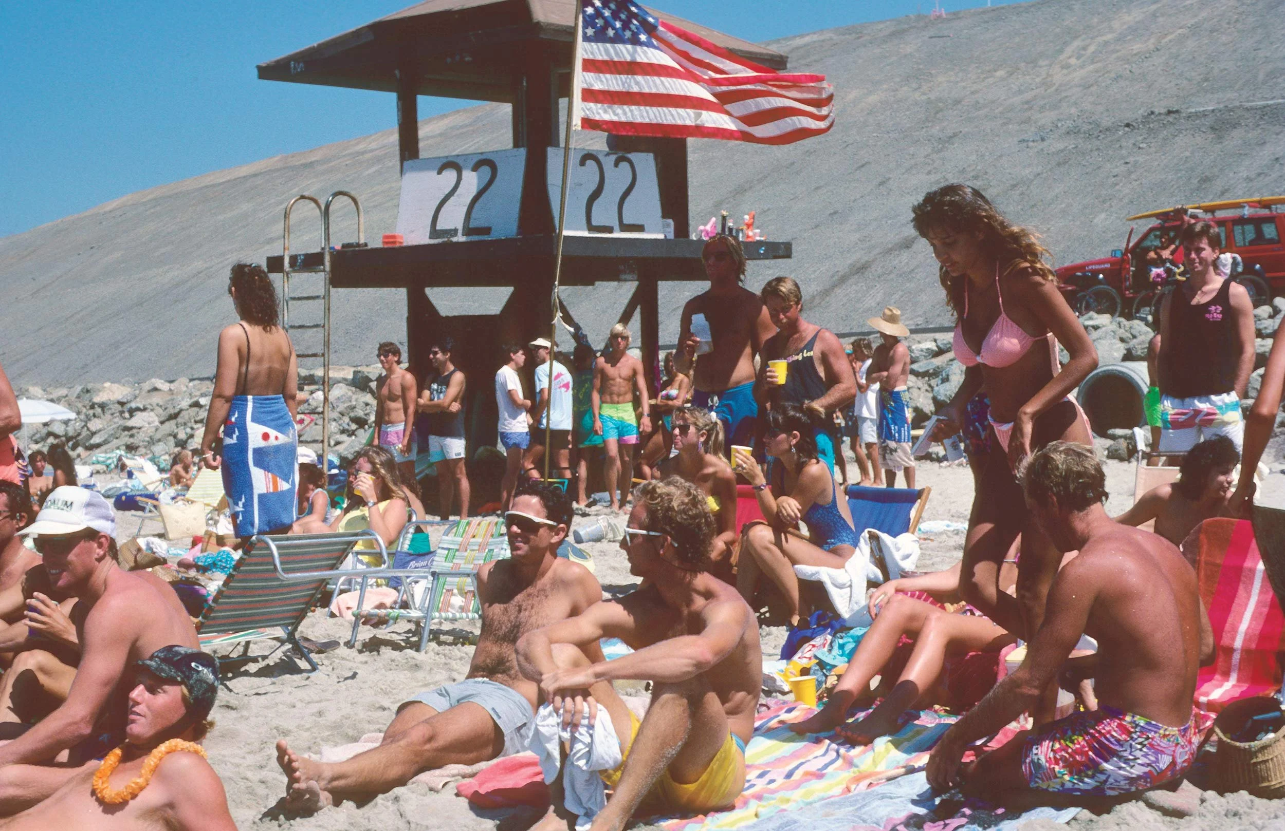 Beach scene with many people, some lounging on chairs and others standing, under a lifeguard tower with an American flag. The background shows a sandy beach and a steep hill.