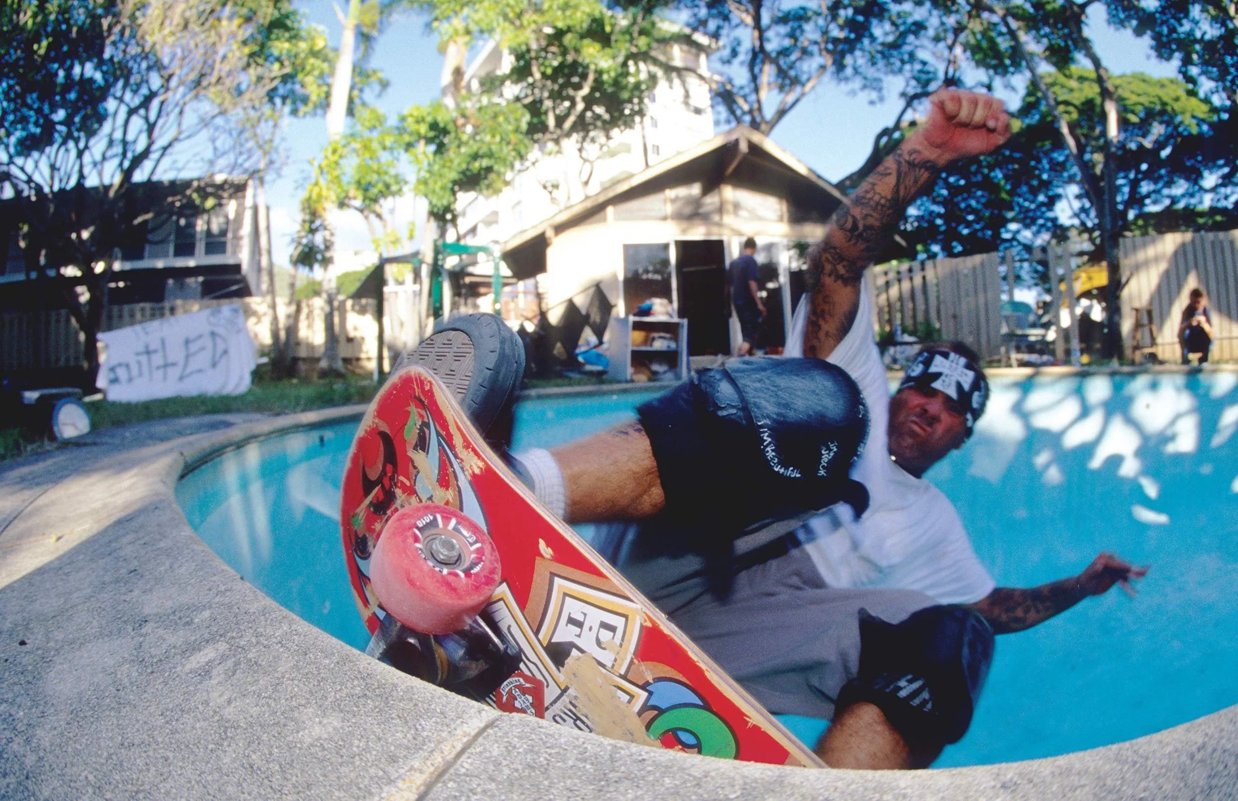 Person with tattoos falling into a swimming pool holding a skateboard with colorful graphics, surrounded by trees and a house in the background.