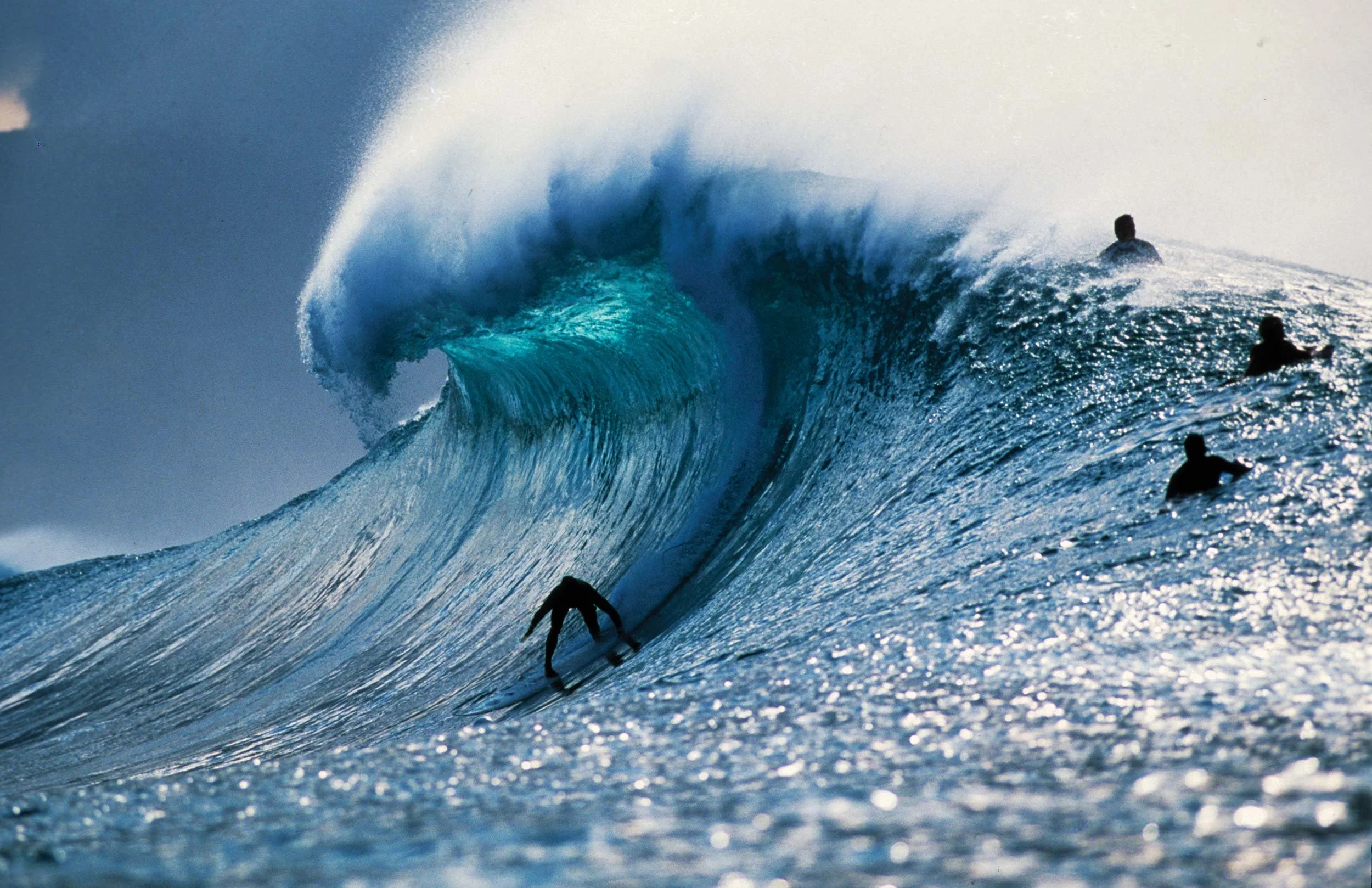 A surfer riding a large ocean wave with others in the water nearby, with the wave cresting and spray in the air.