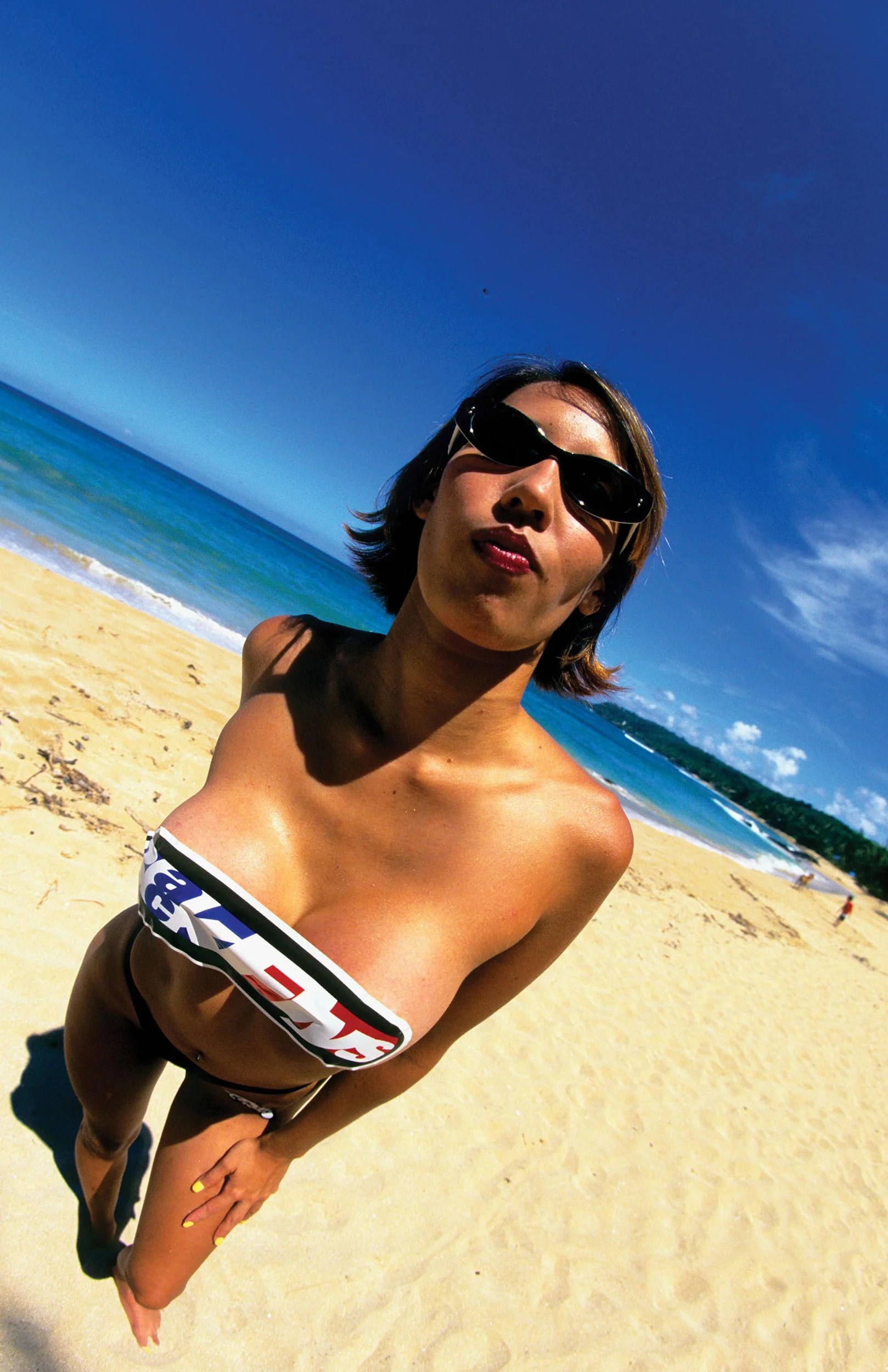 Woman in sunglasses and bikini on beach with sand, ocean, and blue sky in background.