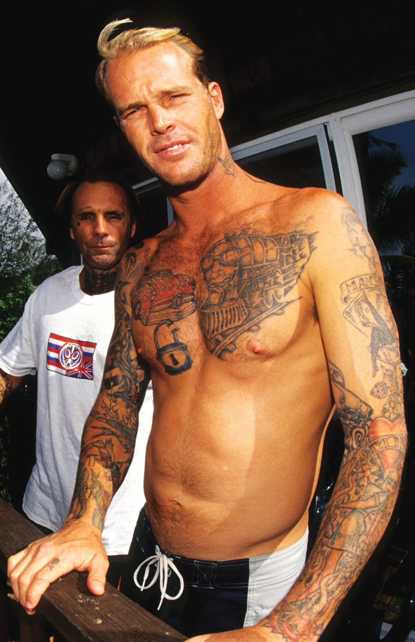A shirtless man with numerous tattoos on his chest and arms standing outdoors with a man in a white T-shirt behind him, near a wooden table or surface, during daylight.
